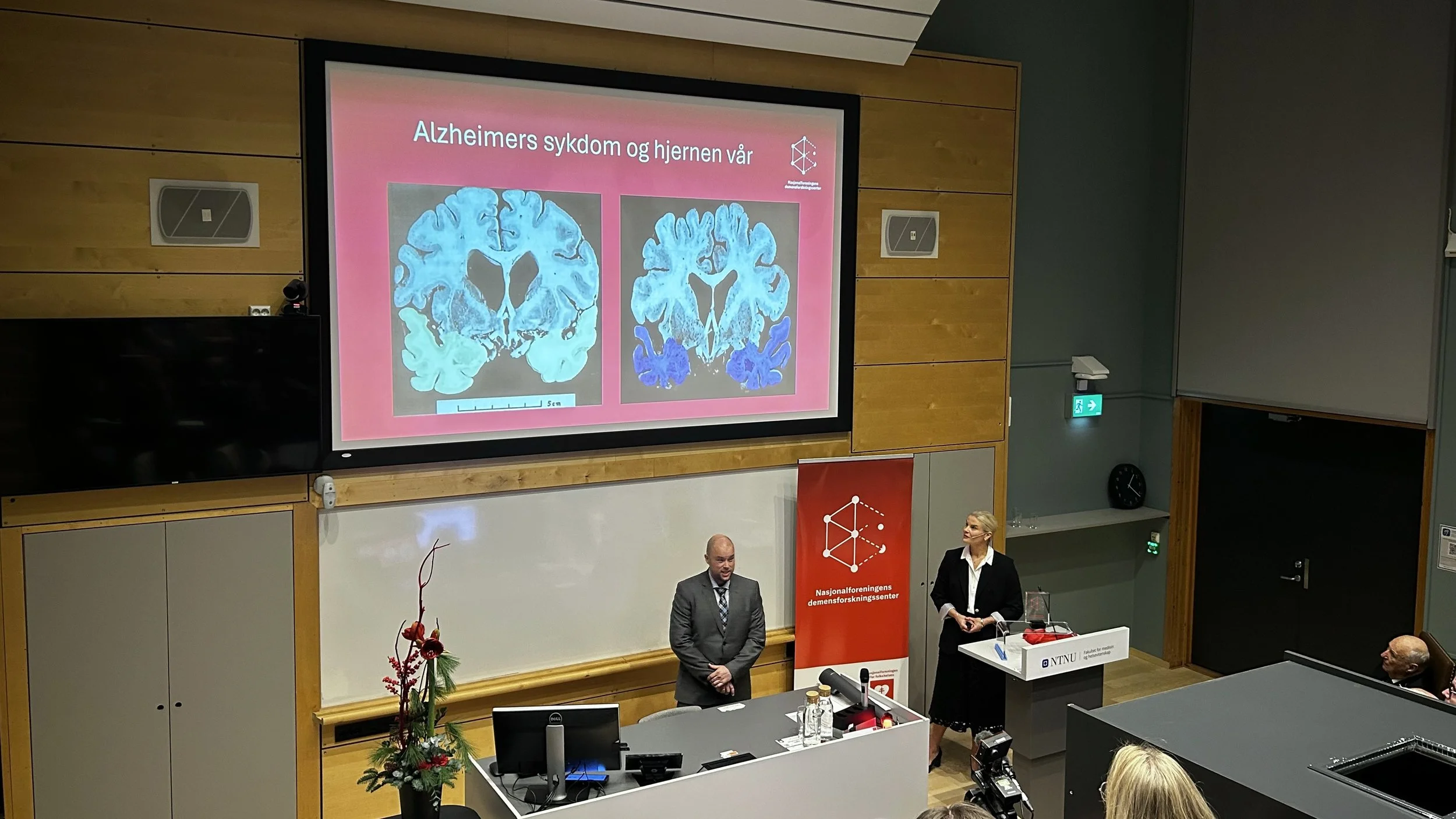 Asgeir Kobro-Flatmoen og Gøril Rolfseng Grøntvedt taler ved podiet i et auditorium. Lysbildet bak dem viser teksten "Alzheimers sykdom og hjernen vår" med bilde av to ulike hjernecanninger, hvor man ser tydelige forskjeller. Fotografi.