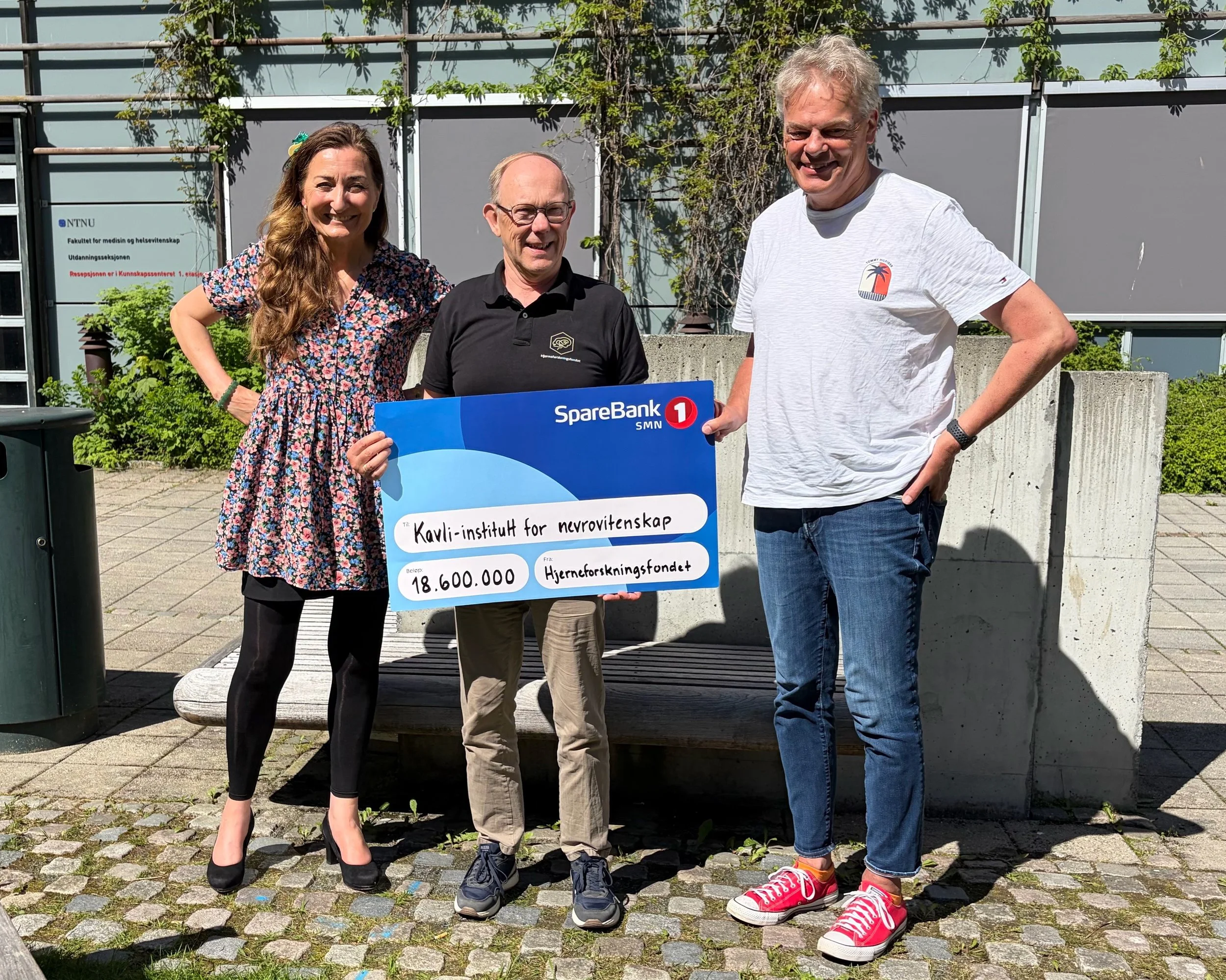 May-Britt Moser, Jan Morten Dyrstad og Edvard Moser smiler mot kamera. Jan Morten Dyrstad holder en sjekk der det fremommer at Hjerneforskningsfondet tildeler Kavli-institutt for nevrovitenskap 18,6 millioner kroner. Foto.