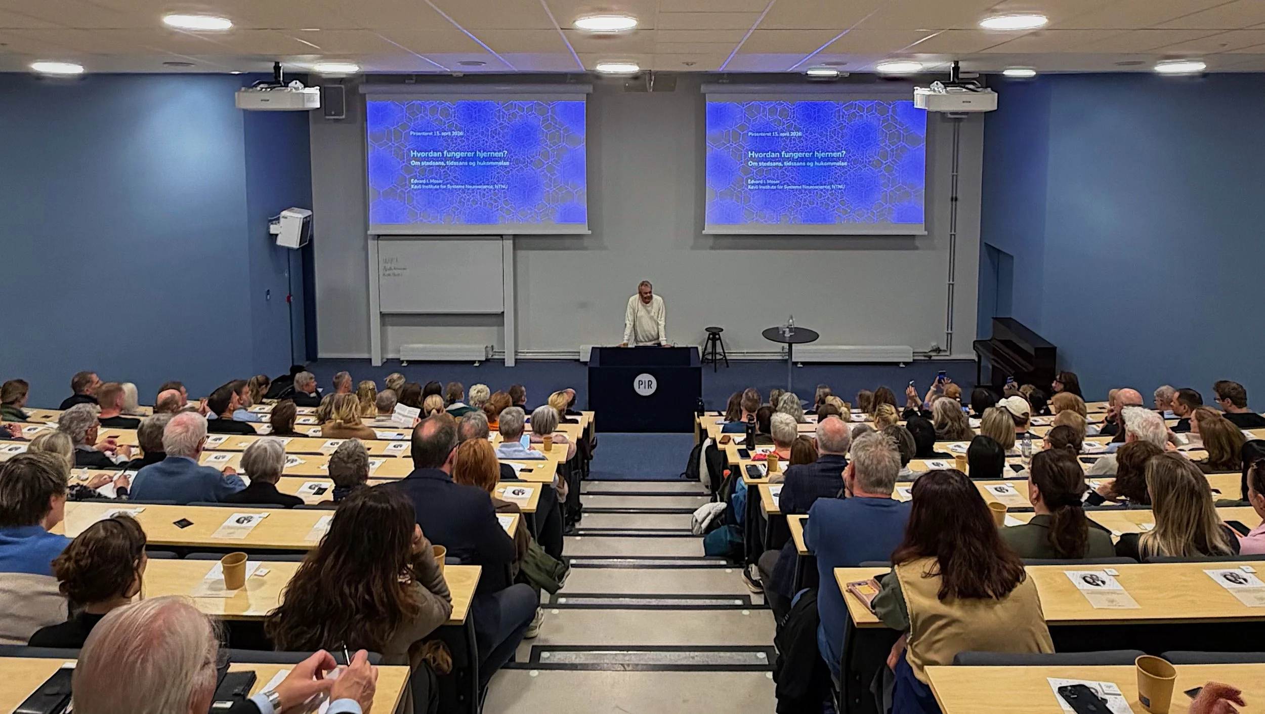 Et fullsatt auditorium, der vi ser et titalls mennesker sittede vendt mot podiet. I front står Edvard Moser og ser ut over forsamlingen. Fotografi.