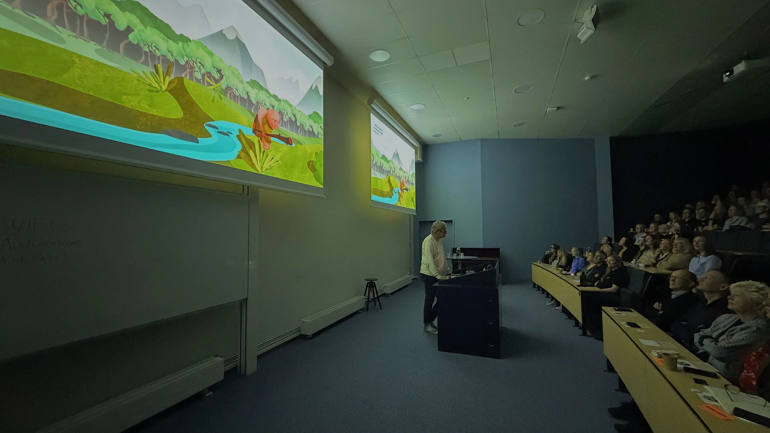 Vi ser podiet i et auditorium fra siden. Ved pulten står Edvard Moser og ser ut mot en forsamling. Forsamlingen ser opp på skjermen, som viser en fargesterk illustrasjon av et urmenneske.