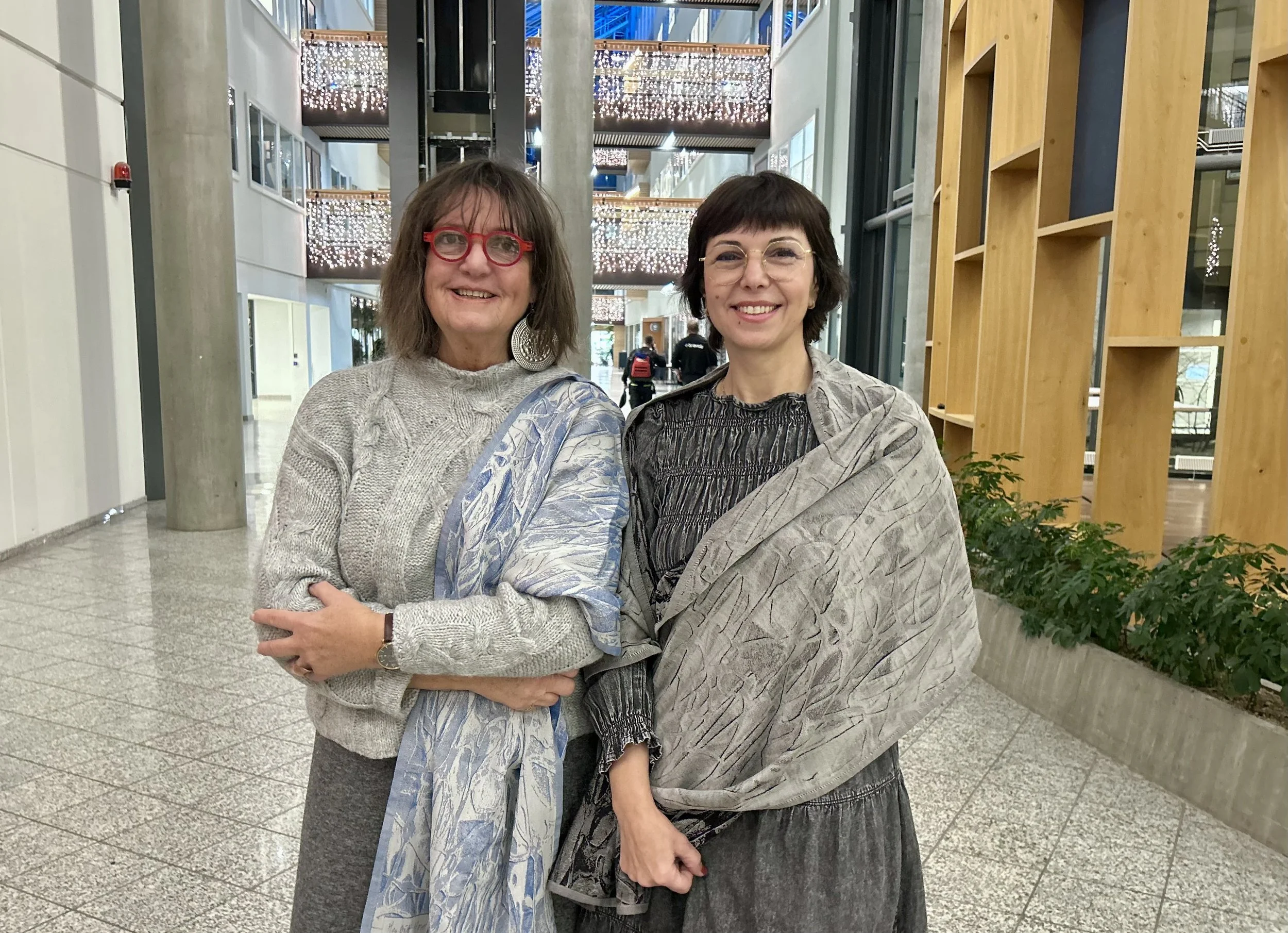 Randi Bakken med blått sjal drapert over den ene skulderen, og Kristina Karlsen med grått skjerf drapert rundt begge skuldre. Begge smiler mot kamera. Fotografi.