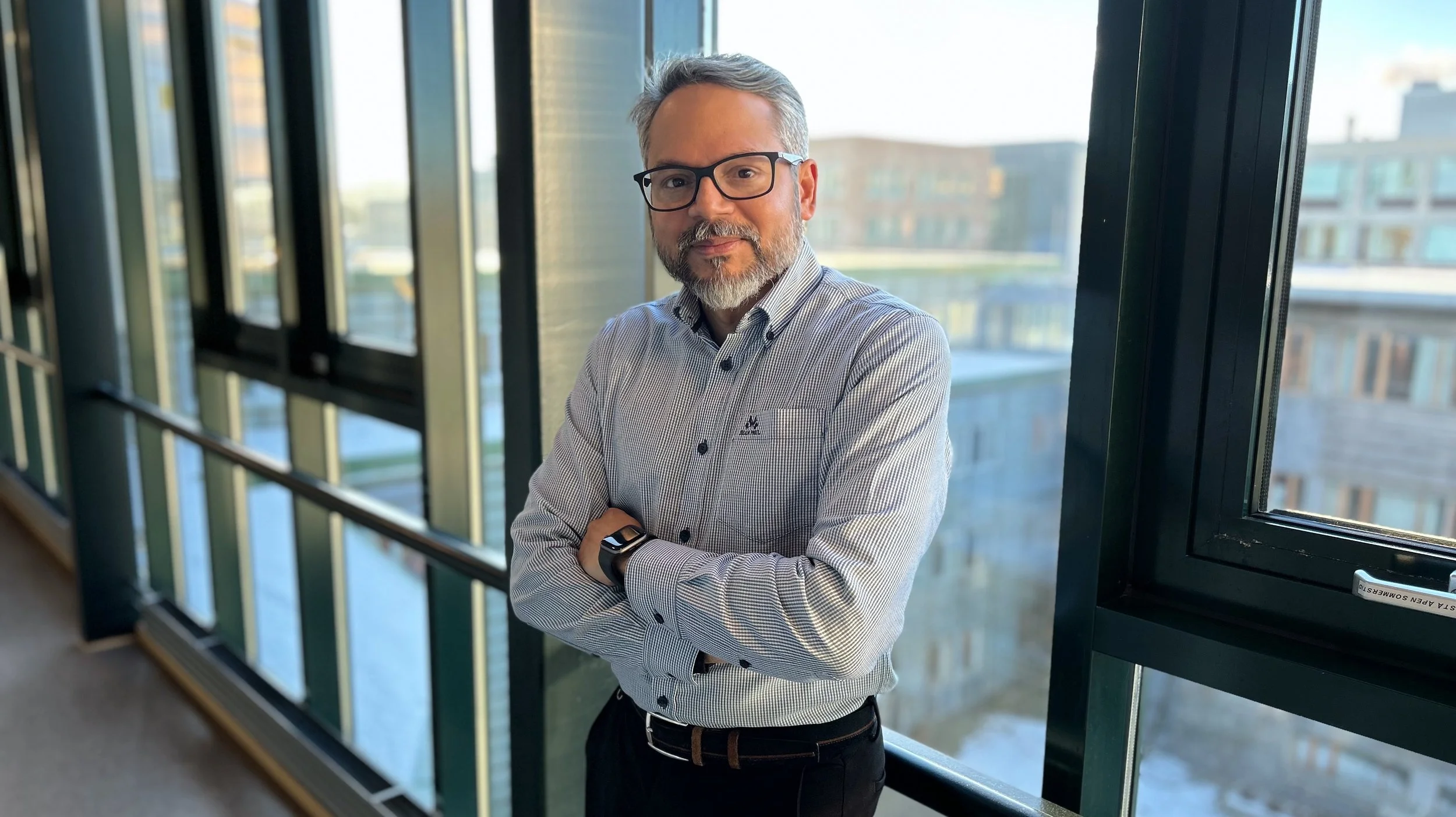 Rajeevkumar Raveendran Nair står lent mot et rekkverk med vinduer ut mot sykehusområdet på St. Olavs hospital. Han har på seg briller, mørk bukse og en stripete skjorte. Han smiler litt mot kamera og har armene korslagt over brystet. Fotografi.