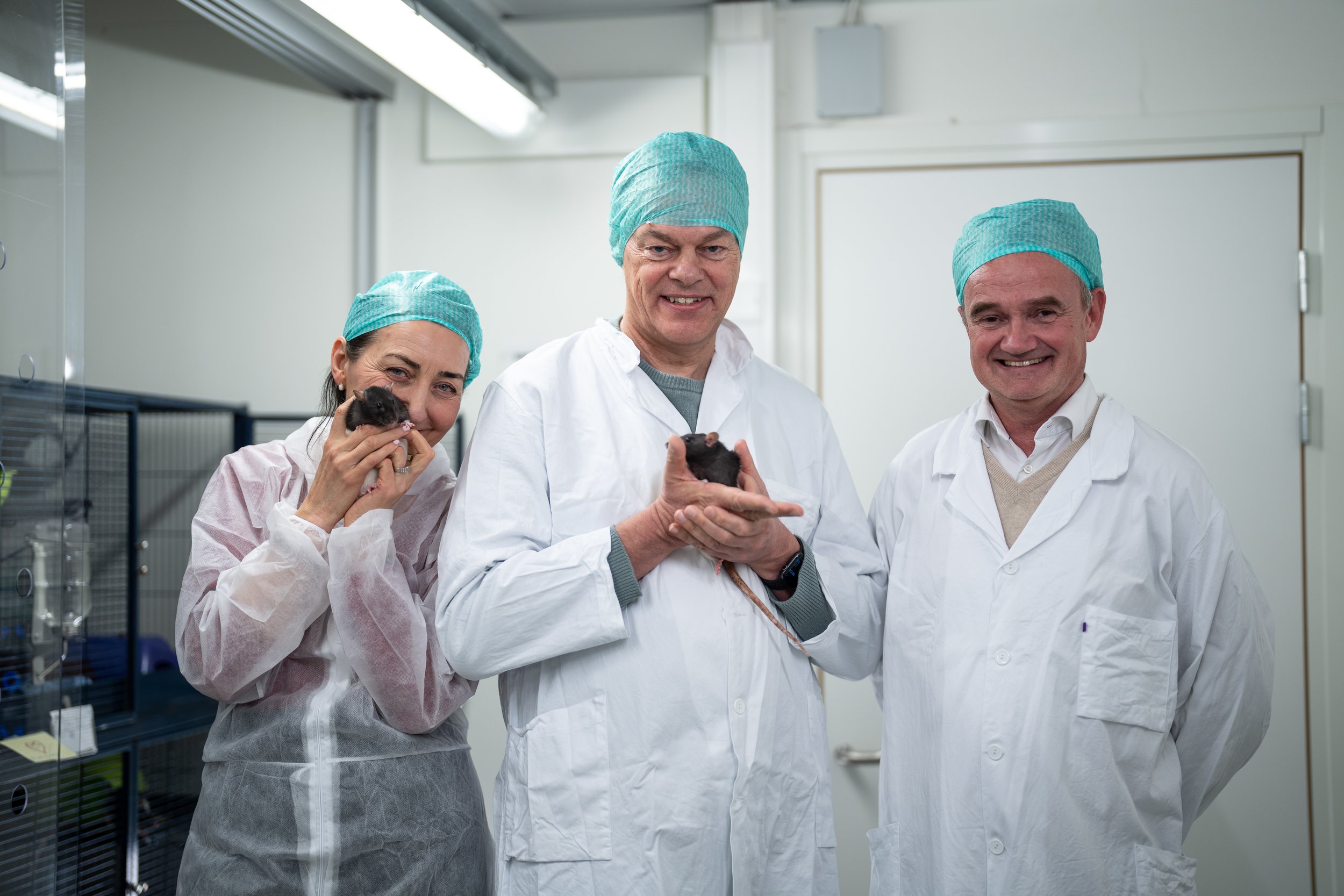 May-Britt Moser, Edvard Moser og Jan-Frode Janson avbildet på dyrestallen. Alle er ikledd labfrakker/overtrekkstøy og hetter. May-Britt og Edvard holder hver sin rotte. Alle smiler mot kamera. Fotografi.