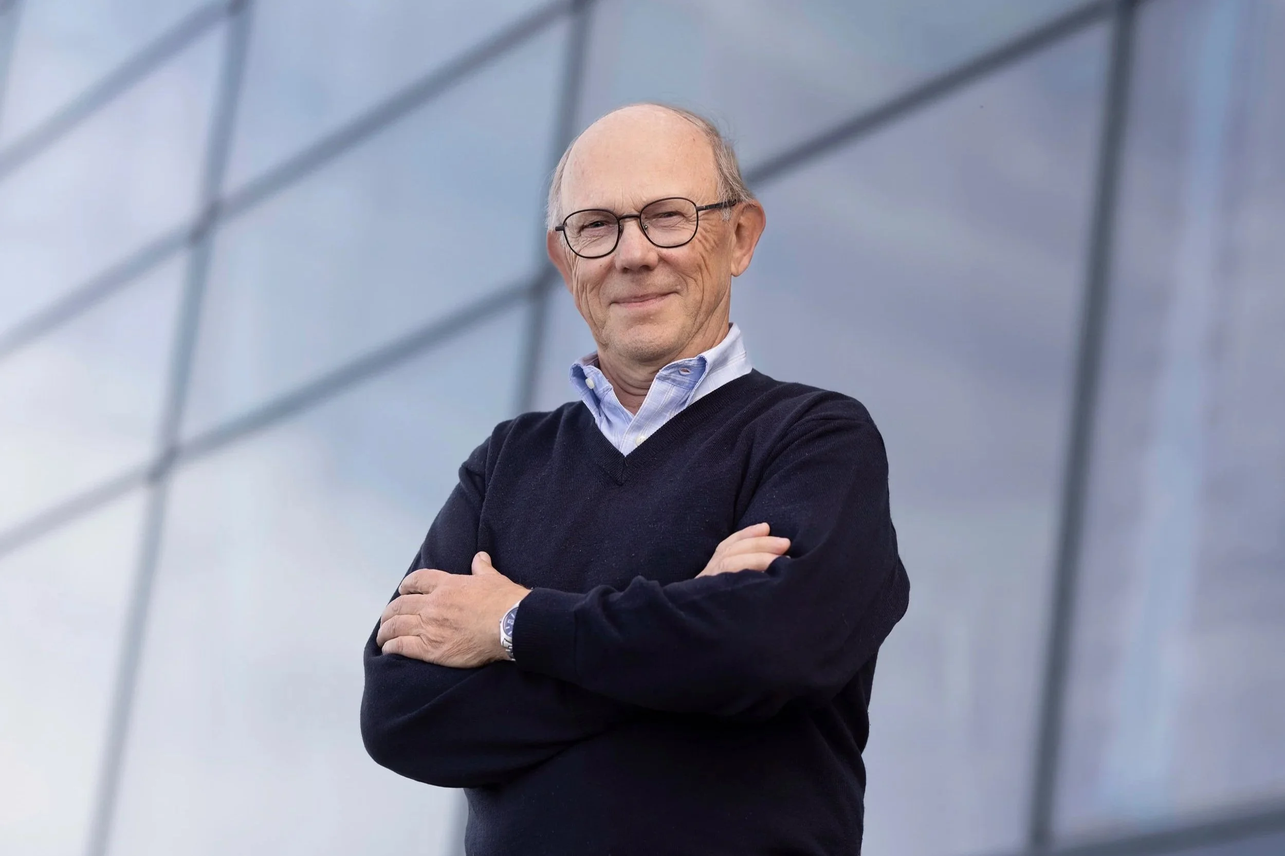 Portrettbilde av Jan Morten Dyrstad. Han er ikledd mørkeblå genser med blå skjorte under, og har armene i kors over brystet. Han smiler mot kamera. Fotografi.