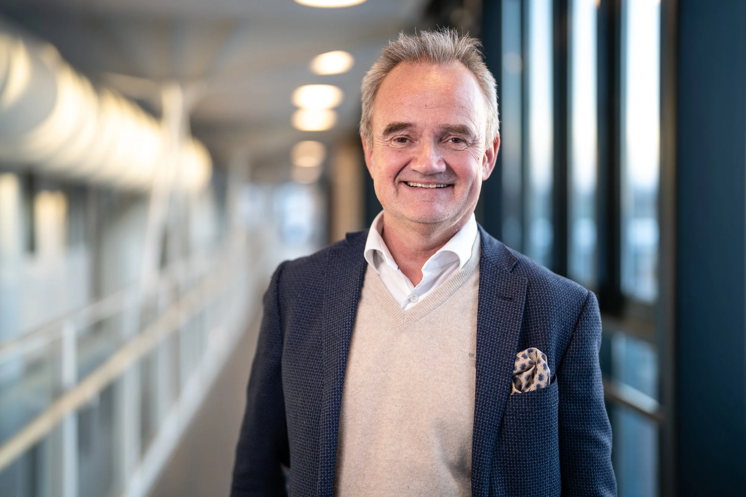 Portrettbilde av Jan-Frode Janson. Han er ikledd blå, rutete blazer og beige genser med hvit skjorte under. Han smiler mot kamera. Fotografi.