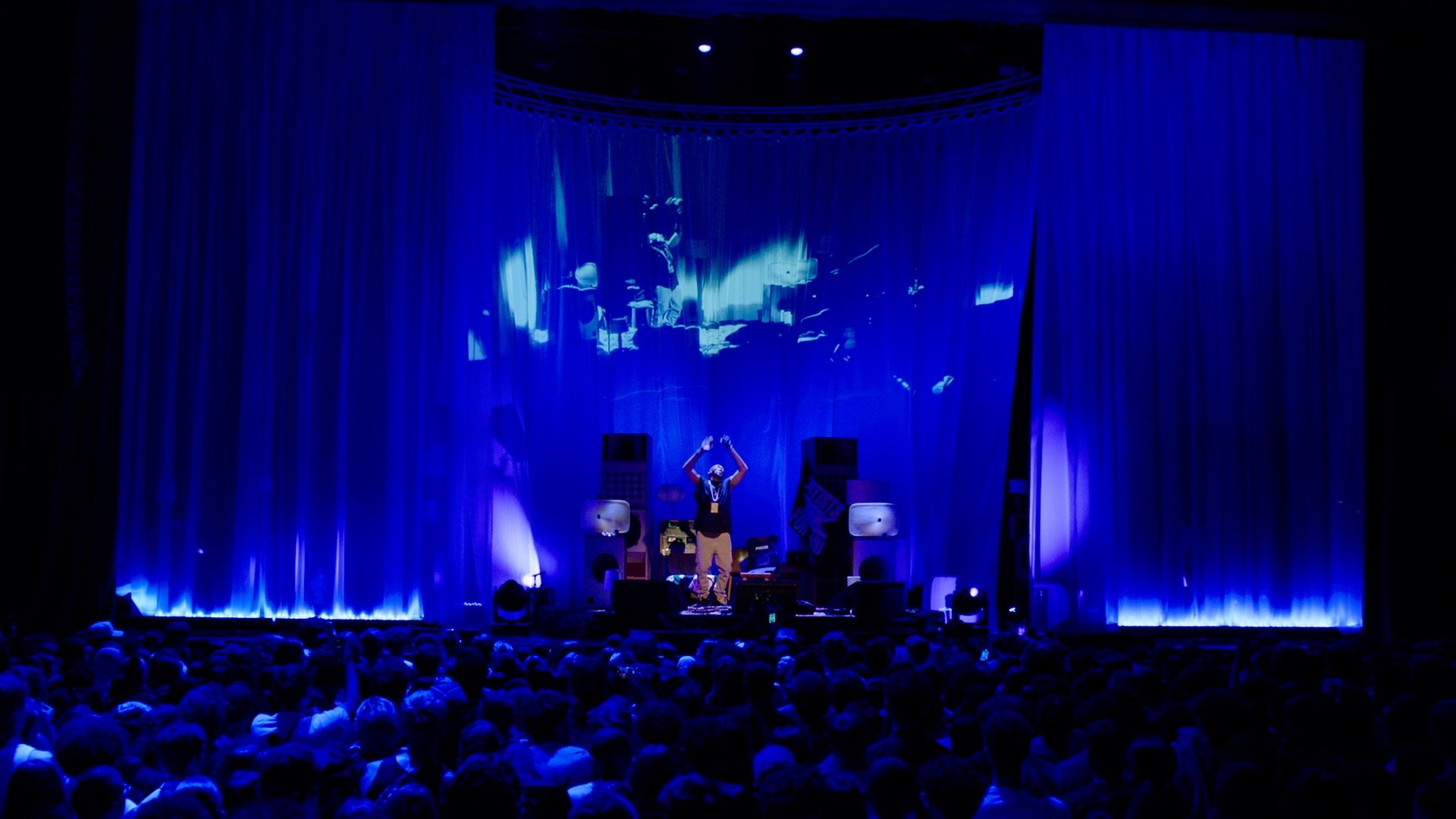 A performer stands on stage under blue lighting, surrounded by sound equipment and a large curtain, with an audience in front.