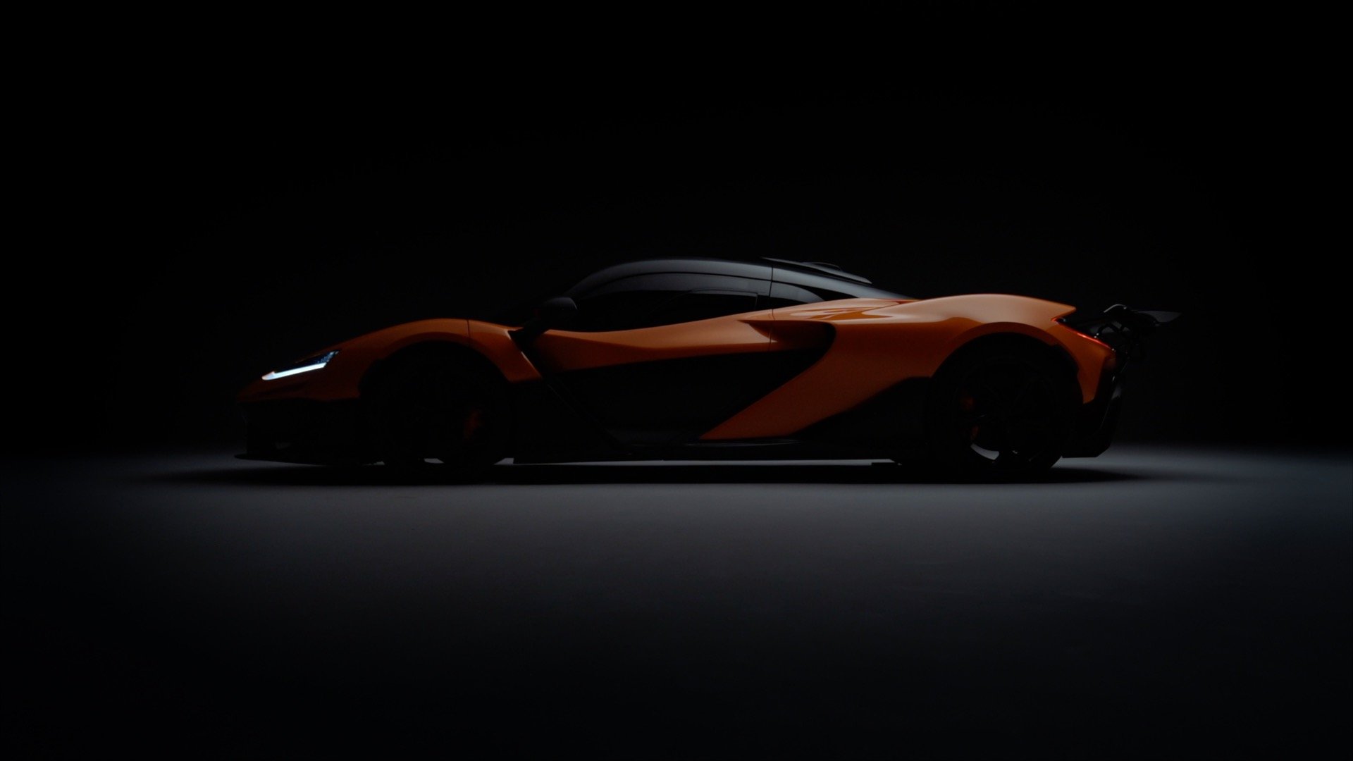 A sleek, orange sports car with black accents, positioned in a dark, minimalistic setting with spotlight highlighting its side profile.