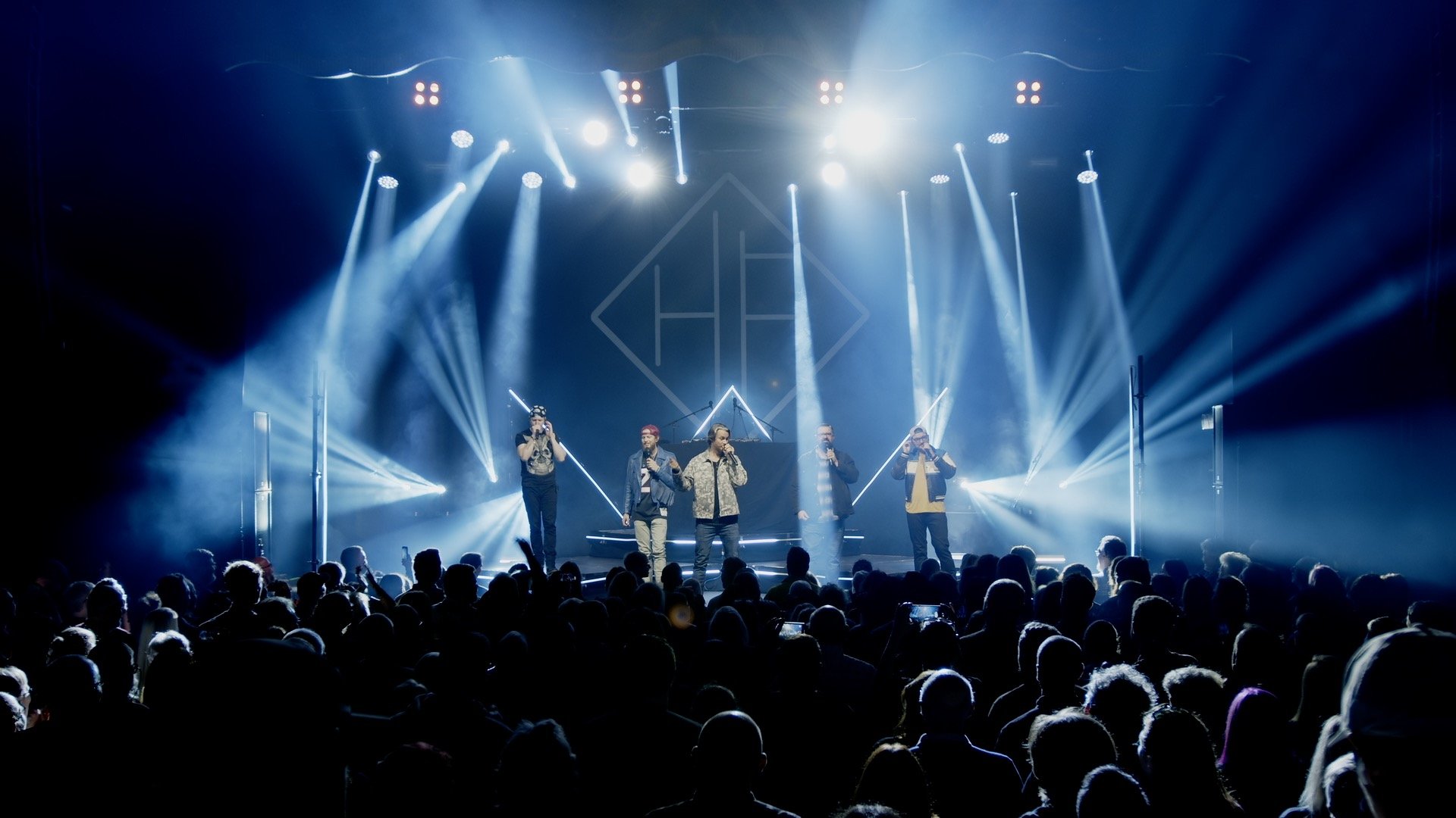 A video camera records a group of young singers performing or rehearsing indoors near large windows, with some audience seating in the foreground.