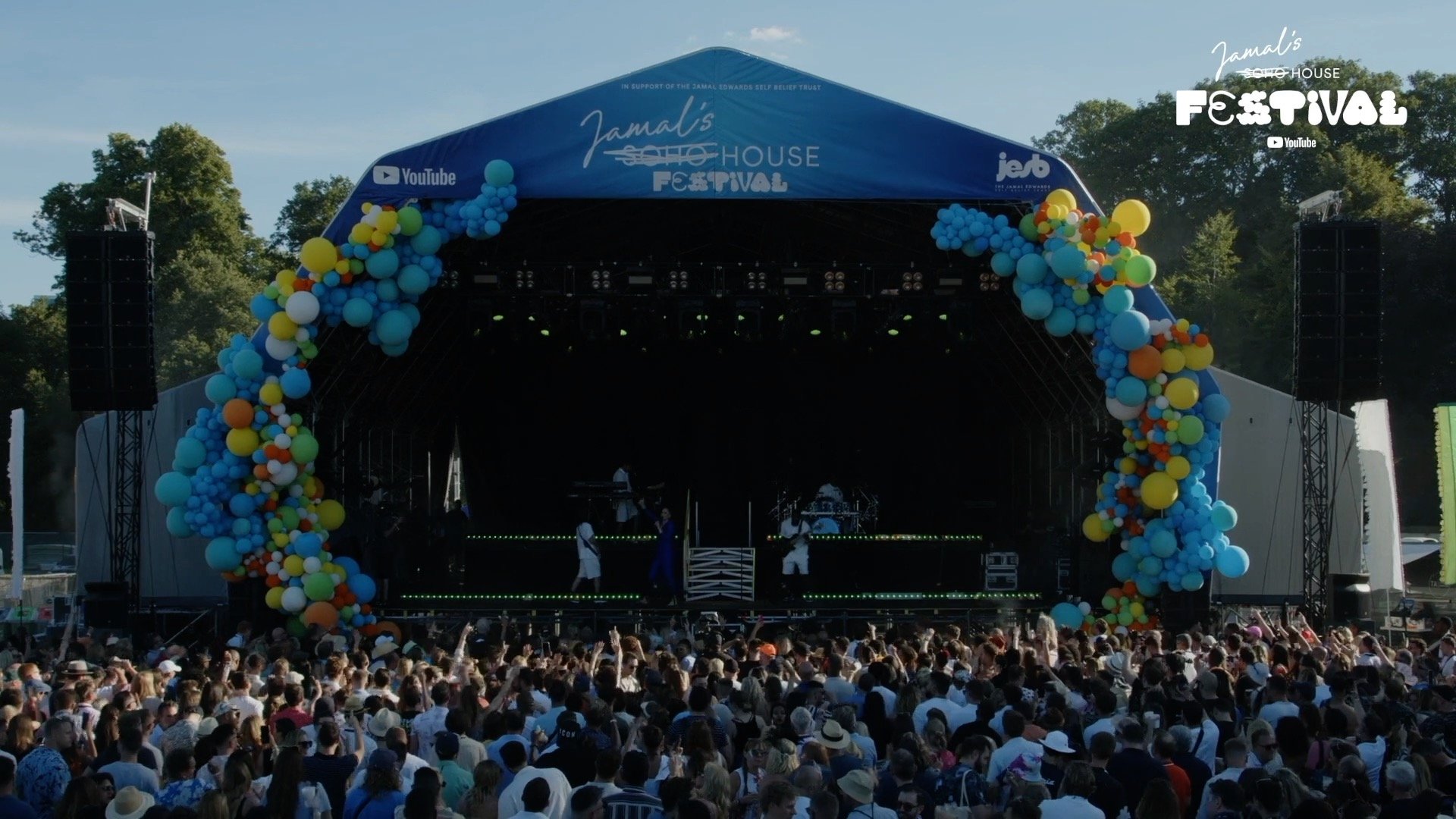 Concert stage decorated with blue, yellow, orange, white, and green balloons, with a large crowd gathered in front, outdoors with trees in the background.