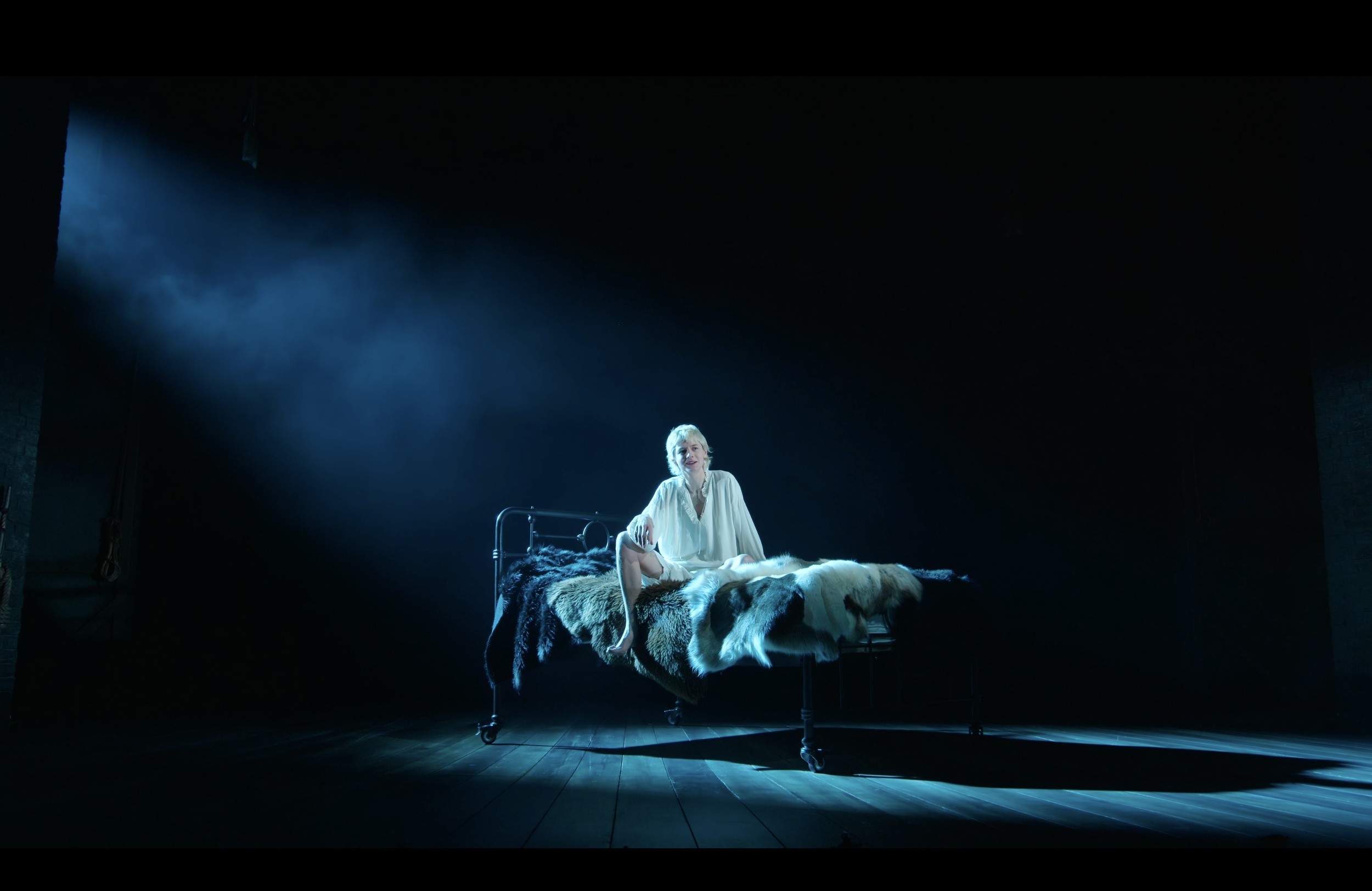 A woman in a white nightgown sitting on a bed with a fur blanket in a dark room illuminated by a single beam of light.