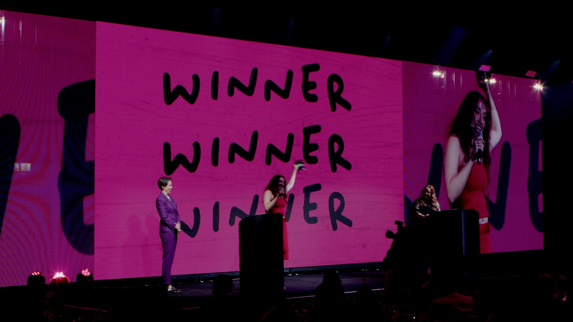 A woman in a red dress holding a microphone on stage with a large screen behind her displaying the words "WINNER" multiple times. She is raising her hand in victory, and another woman in a purple suit is standing nearby on stage.