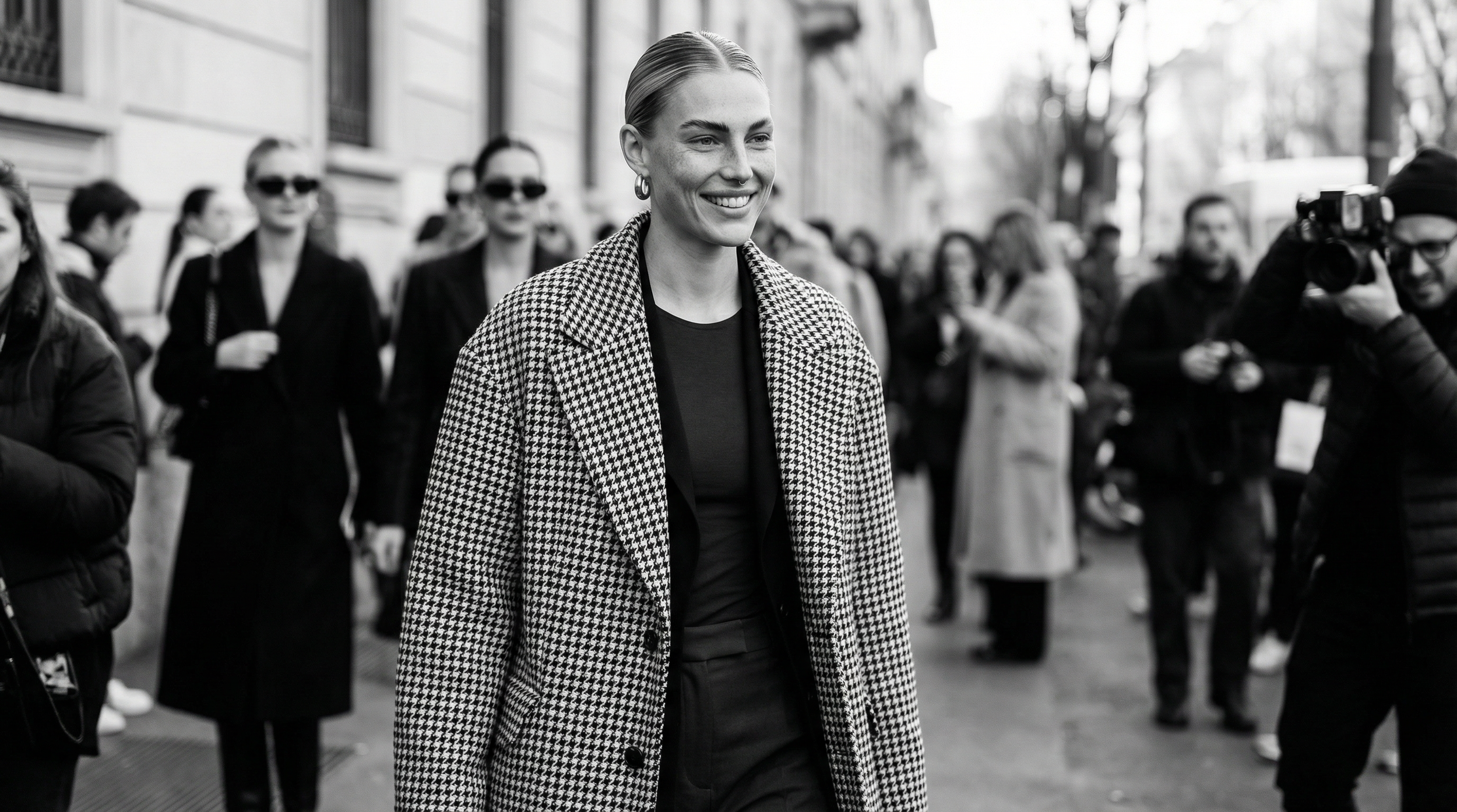 A woman smiling walks on a city street surrounded by people, some with cameras, in black-and-white photo.