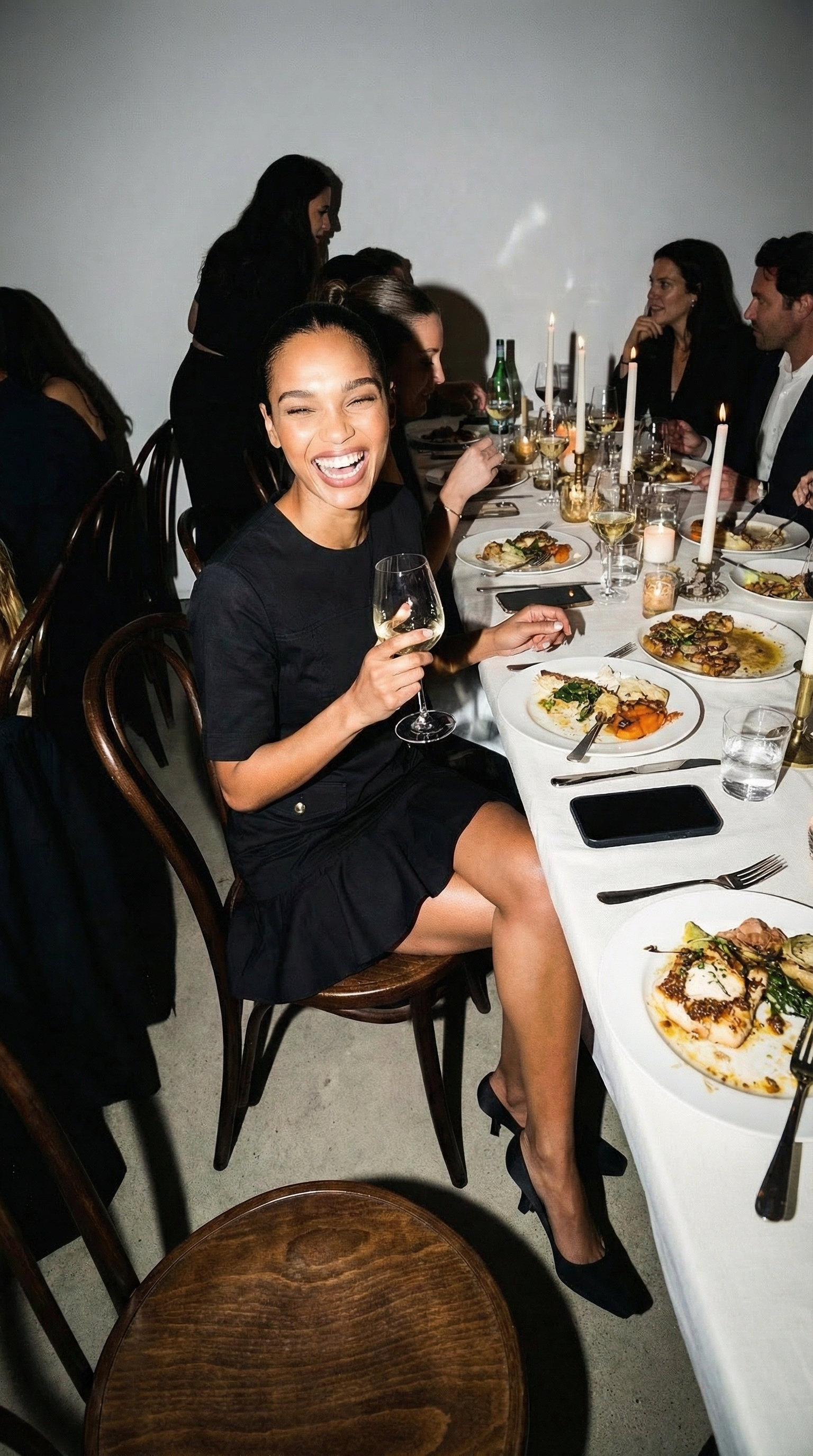 A woman in a black dress sitting at a dinner table, smiling and holding a glass of white wine, with other people dining in the background.