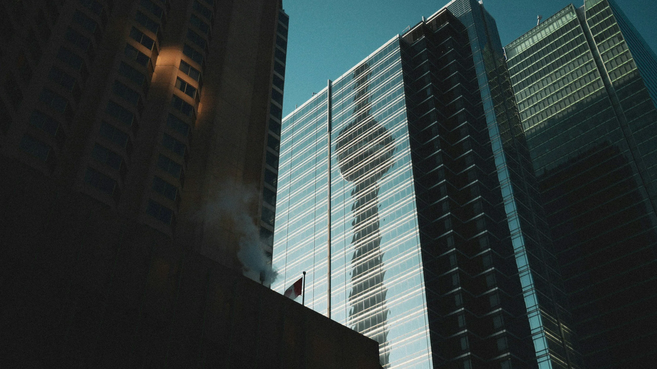 Reflection of a modern skyscraper with glass windows and lines of light, seen in a taller building's glass facade, with other high-rise buildings around.