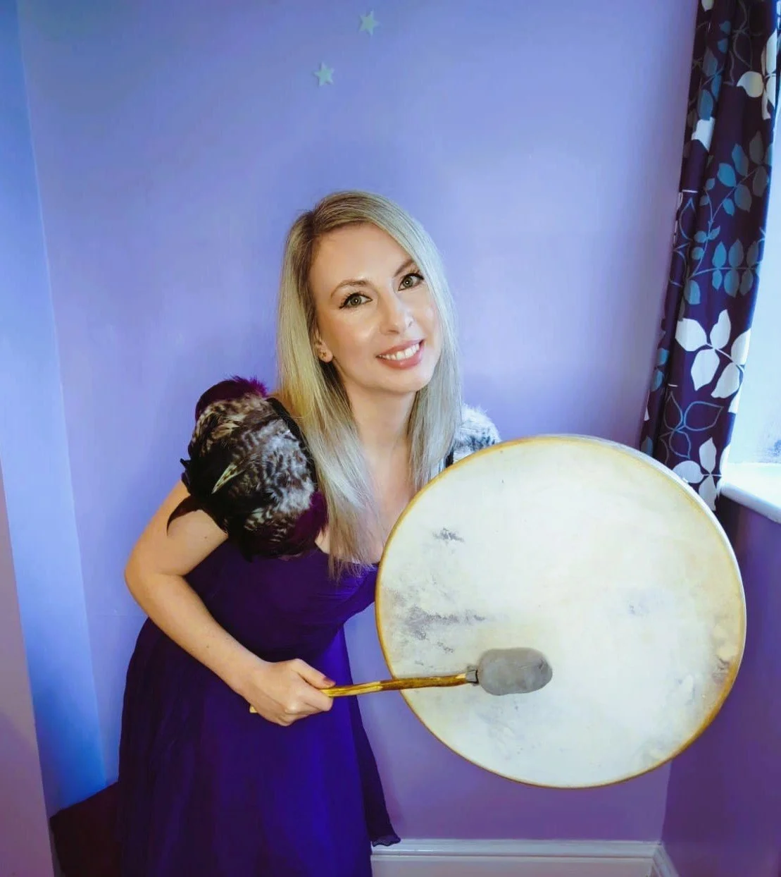 Smiling Pandora Ariel with silver hair stands against a soft purple wall with small stars, holding a round frame drum.Wearing a ceremonial flowing purple dress with feathered shoulders. A High Priestess that uses drumming and immersive sounds.