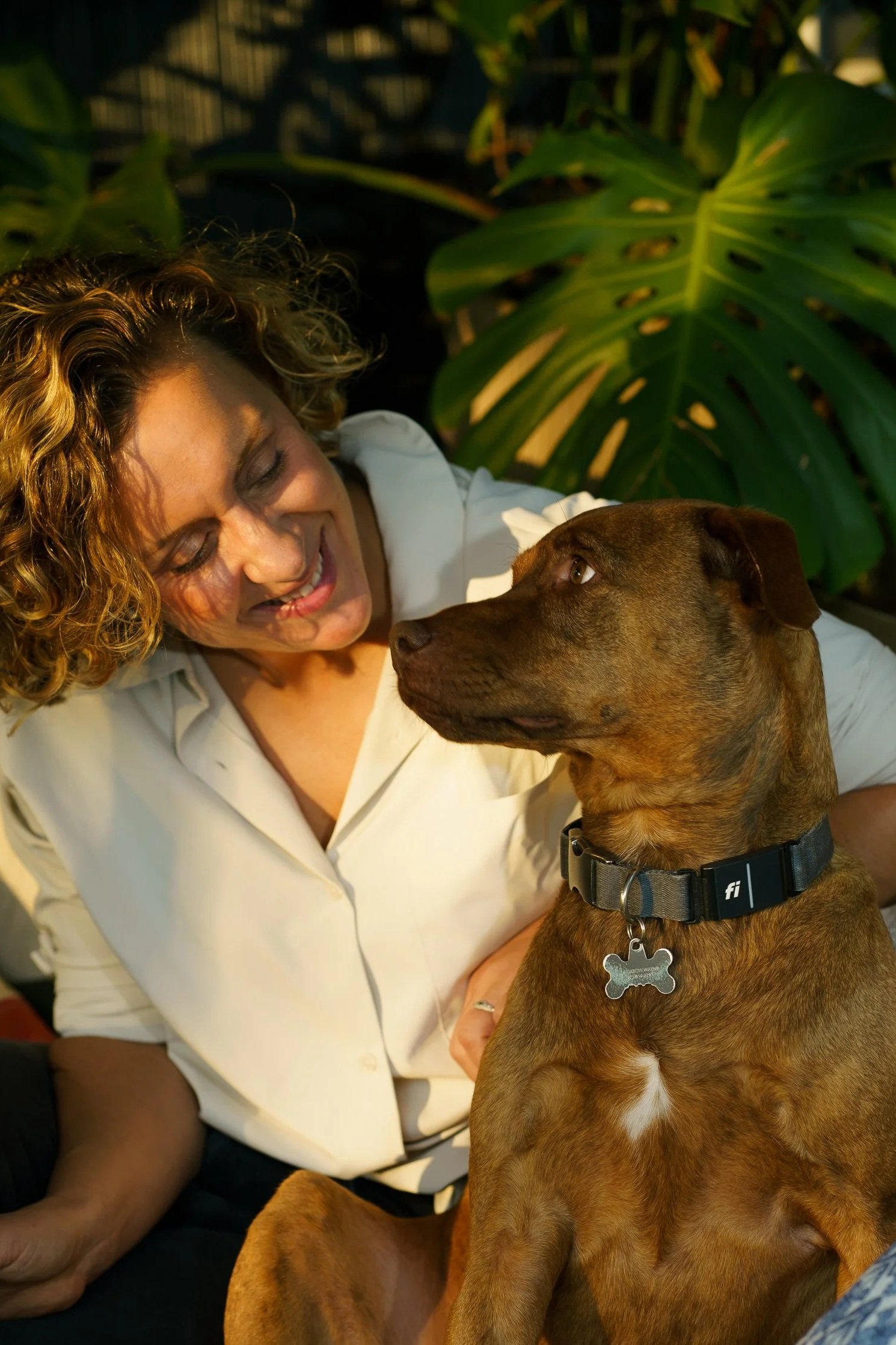 A woman with curly hair smiling and looking at a brown dog with a black collar, sitting next to her, near large green leaves.