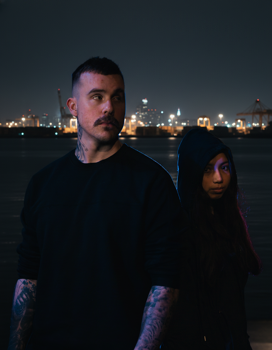 A man and a woman stand outdoors at night with a city skyline and harbor in the background. The man has tattoos and a short haircut, while the woman is wearing a hooded jacket.