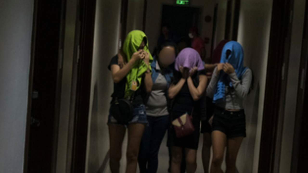 Four young women with colorful headscarves covering their faces standing in an elevator, with people in the background.