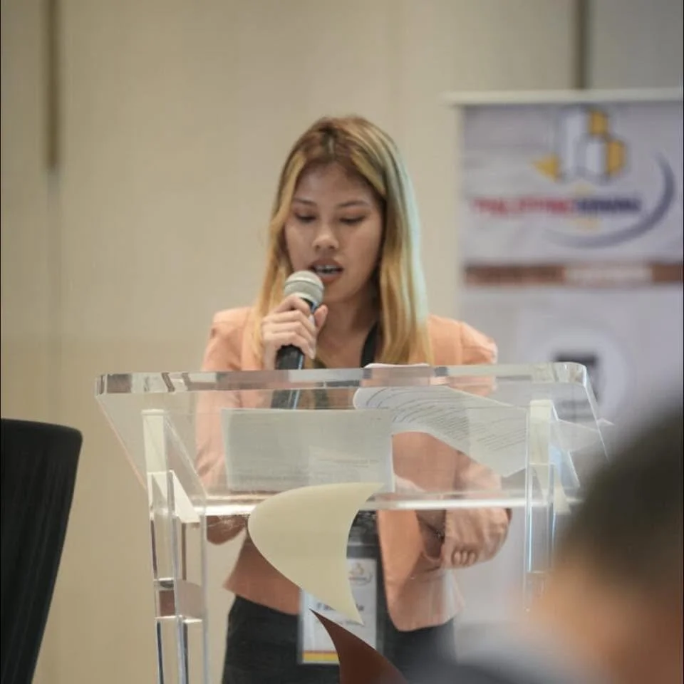 A woman with blonde hair, wearing a light pink blazer, is speaking into a microphone while standing behind a clear acrylic podium that has papers and documents on it. There is a banner in the background.