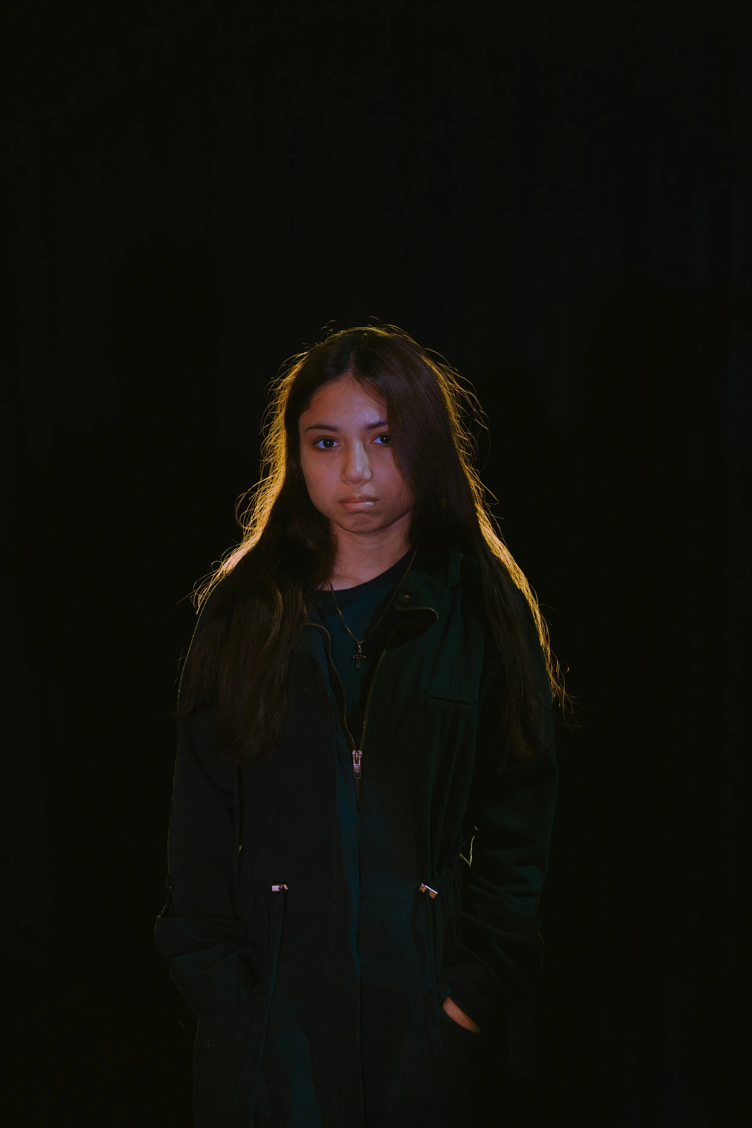 A young girl with long dark hair standing against a black background, wearing a black jacket and a necklace with a cross.