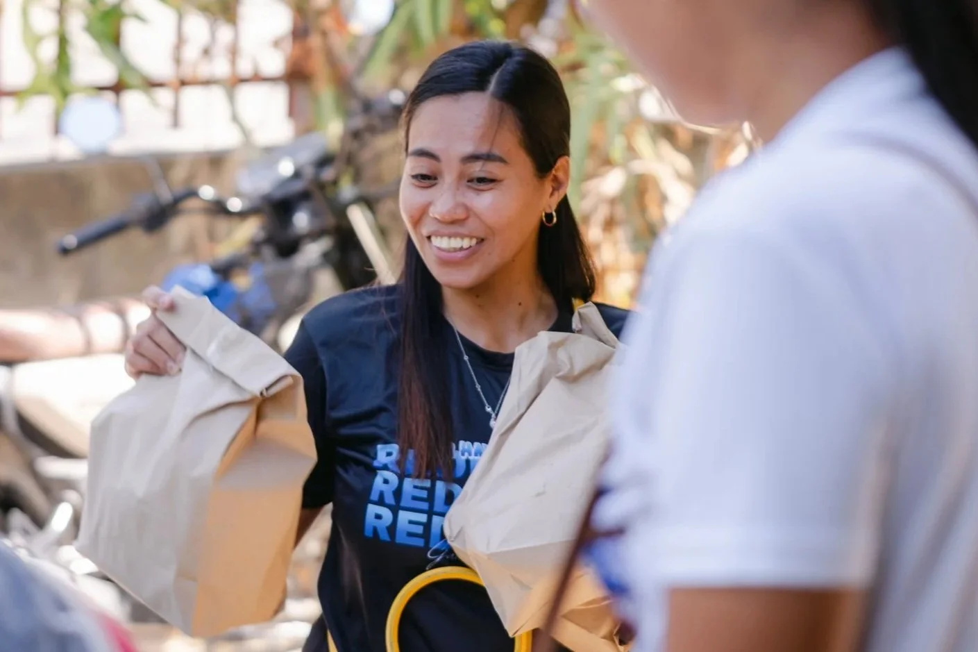 Two women exchanging brown paper bags outdoors, with motorcycles and greenery in background.