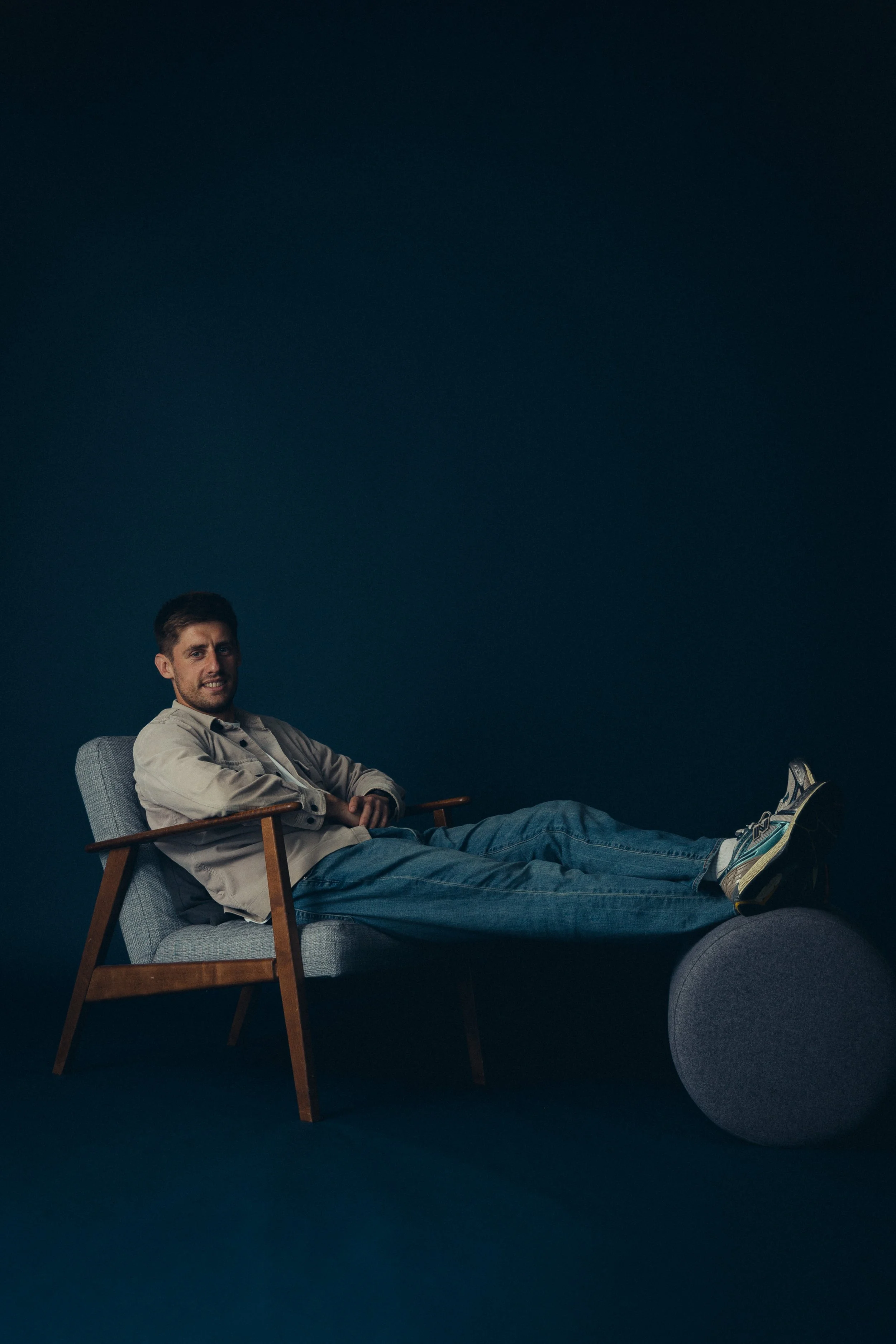 Mischief creative director, Lewis Durkin, sitting in a light gray armchair with wooden armrests, relaxing with his legs extended onto a gray round ottoman, against a dark blue background.