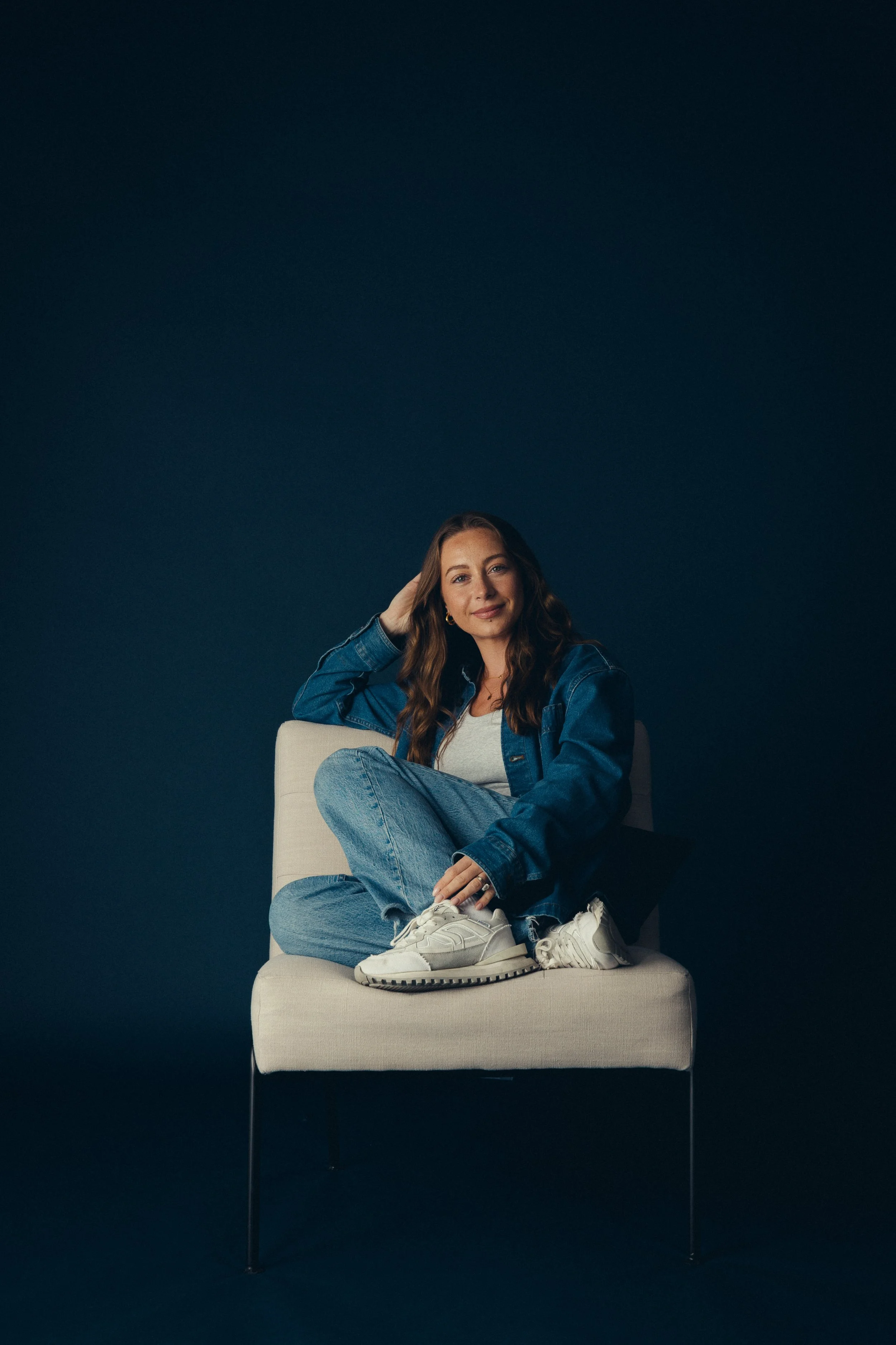 Mischief creative director, Emily Gosen, sitting on a light-colored chair against a dark background, wearing a denim jacket, gray shirt, jeans, and white sneakers, smiling at the camera.