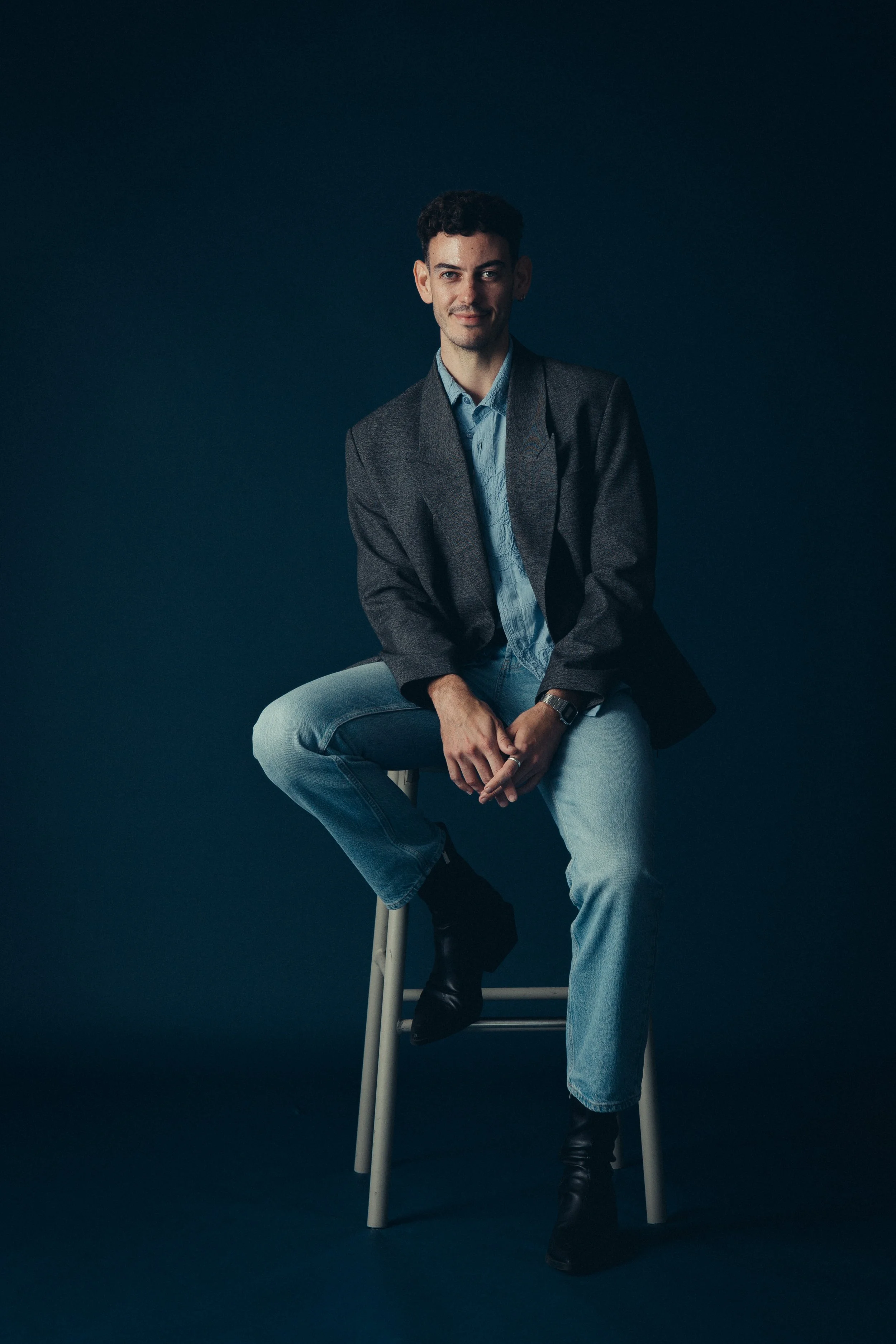 Mischief gen z expert and strategist, Jordan Mulvaney, sitting on a stool against a dark blue background. He is wearing a dark gray blazer, light blue shirt, light blue jeans, and black boots, with a watch on his left wrist.