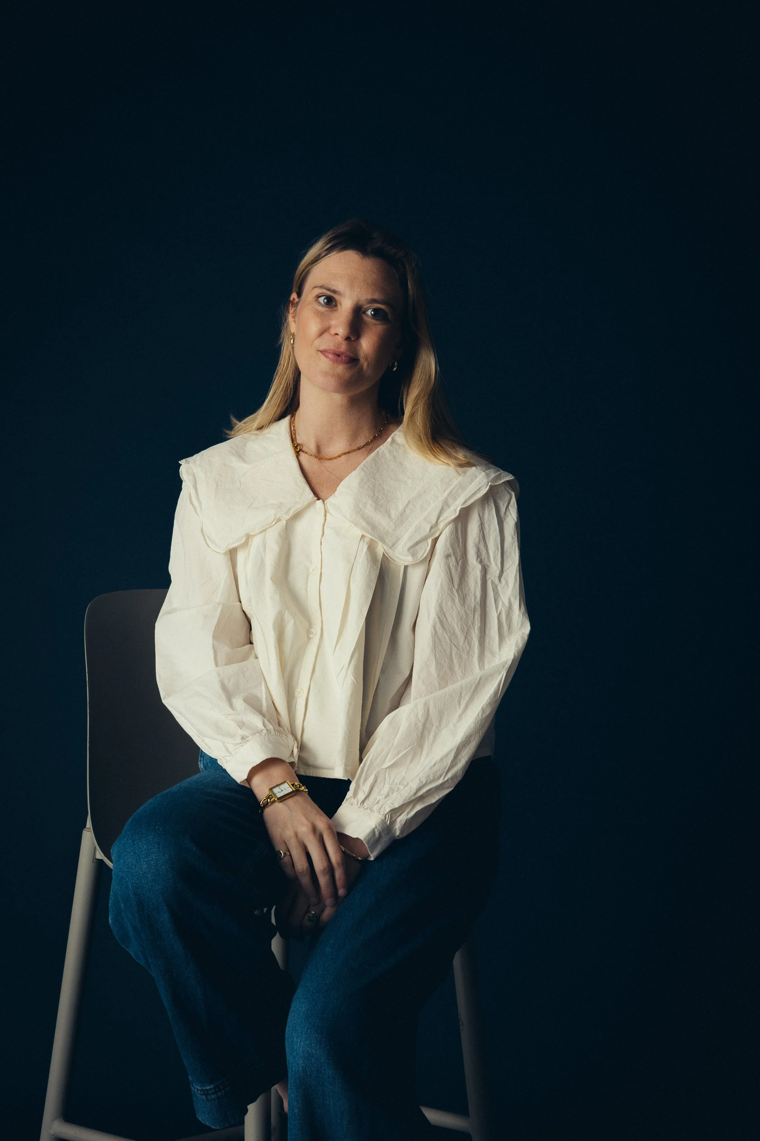 Mischief Creative Director, Ottilie Ross, sitting on a chair against a dark background, wearing a white blouse, jeans, and jewelry.