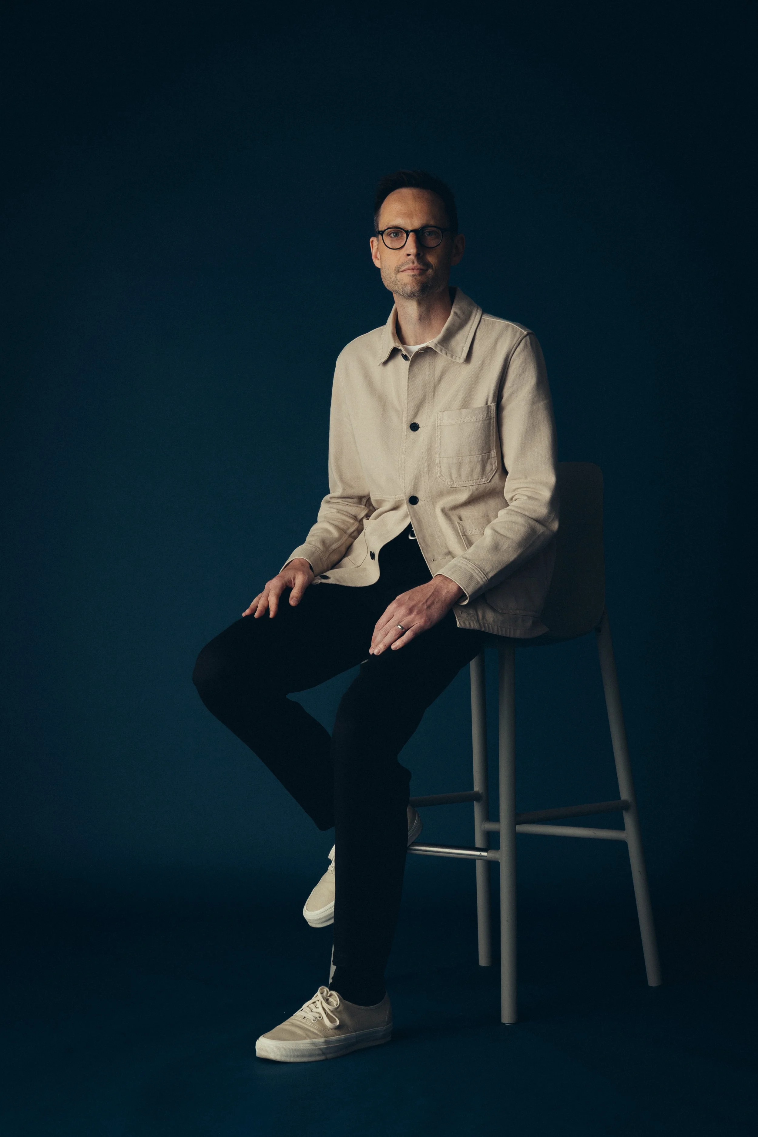 Mischief head of strategy, Dan Deeks-Osburn, wearing glasses, a beige jacket, black pants, and beige sneakers sitting on a stool against a dark background.