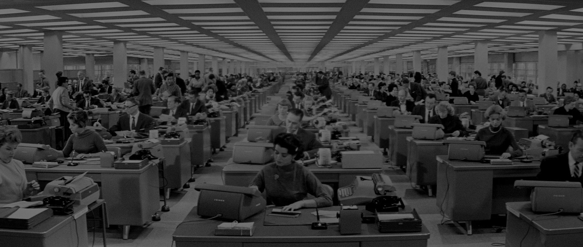 Black and white photo of a busy office with rows of desks, each with a typewriter and telephone, filled with men and women working and talking.