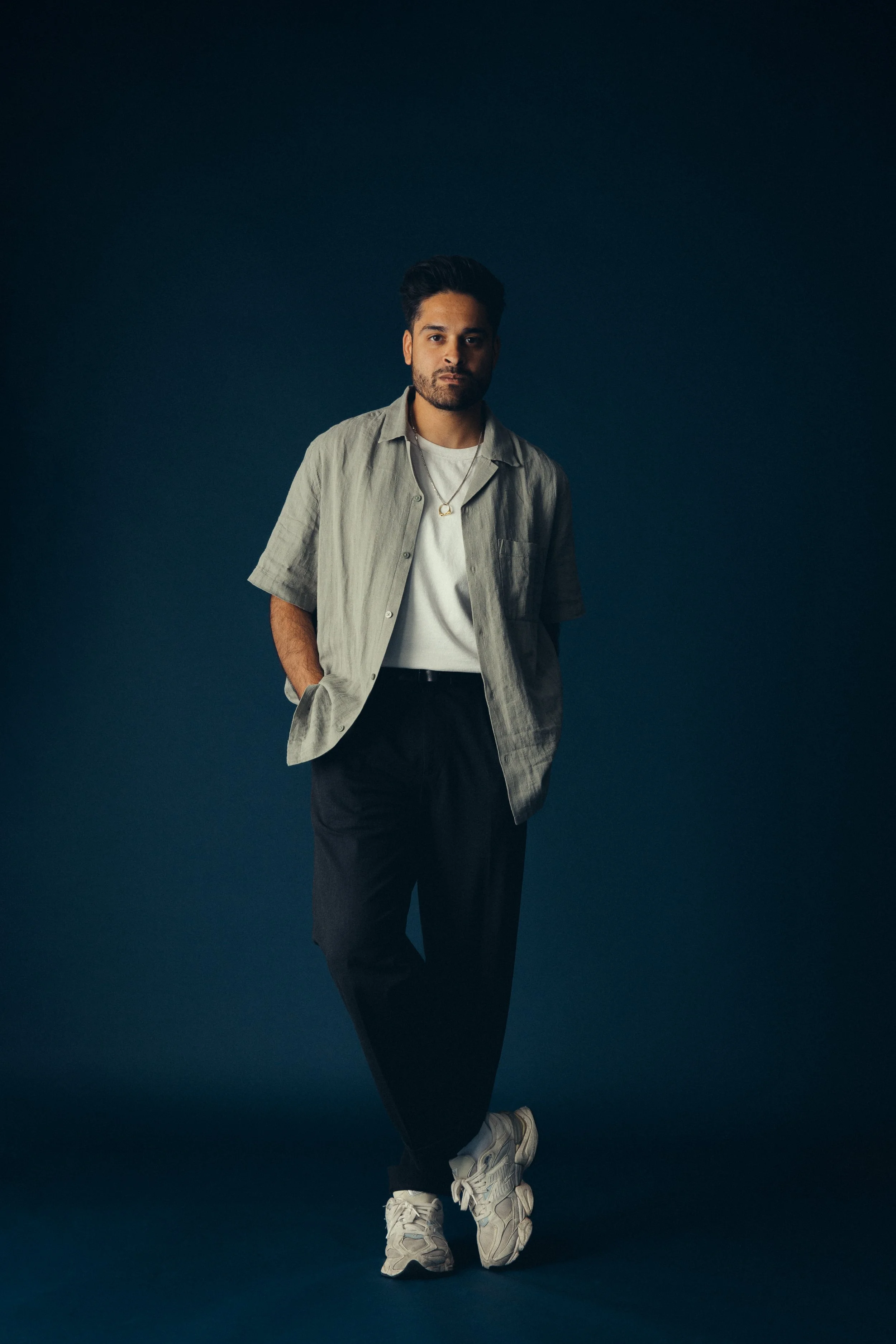 Mischief director, Vish Rana, standing against a dark background, wearing a light gray short-sleeved shirt over a white t-shirt, black pants, and sneakers.
