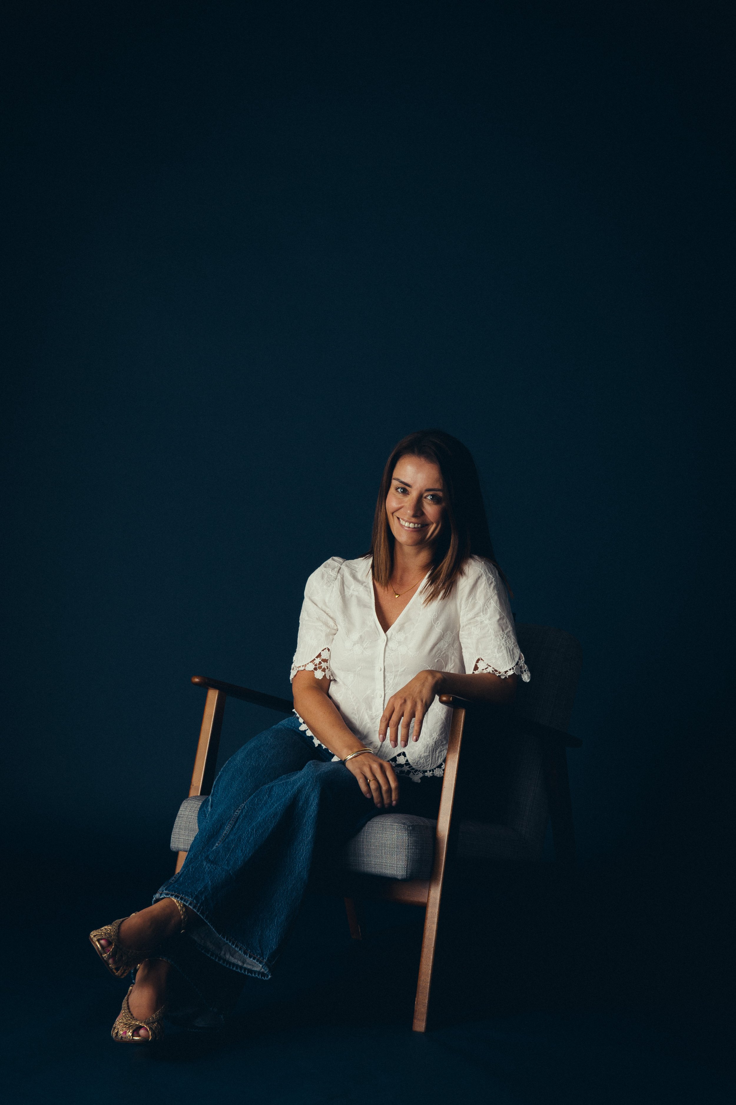 Mischief MD, Charlotte Brooks, sitting in a chair with a dark background, smiling and wearing a white blouse and jeans.