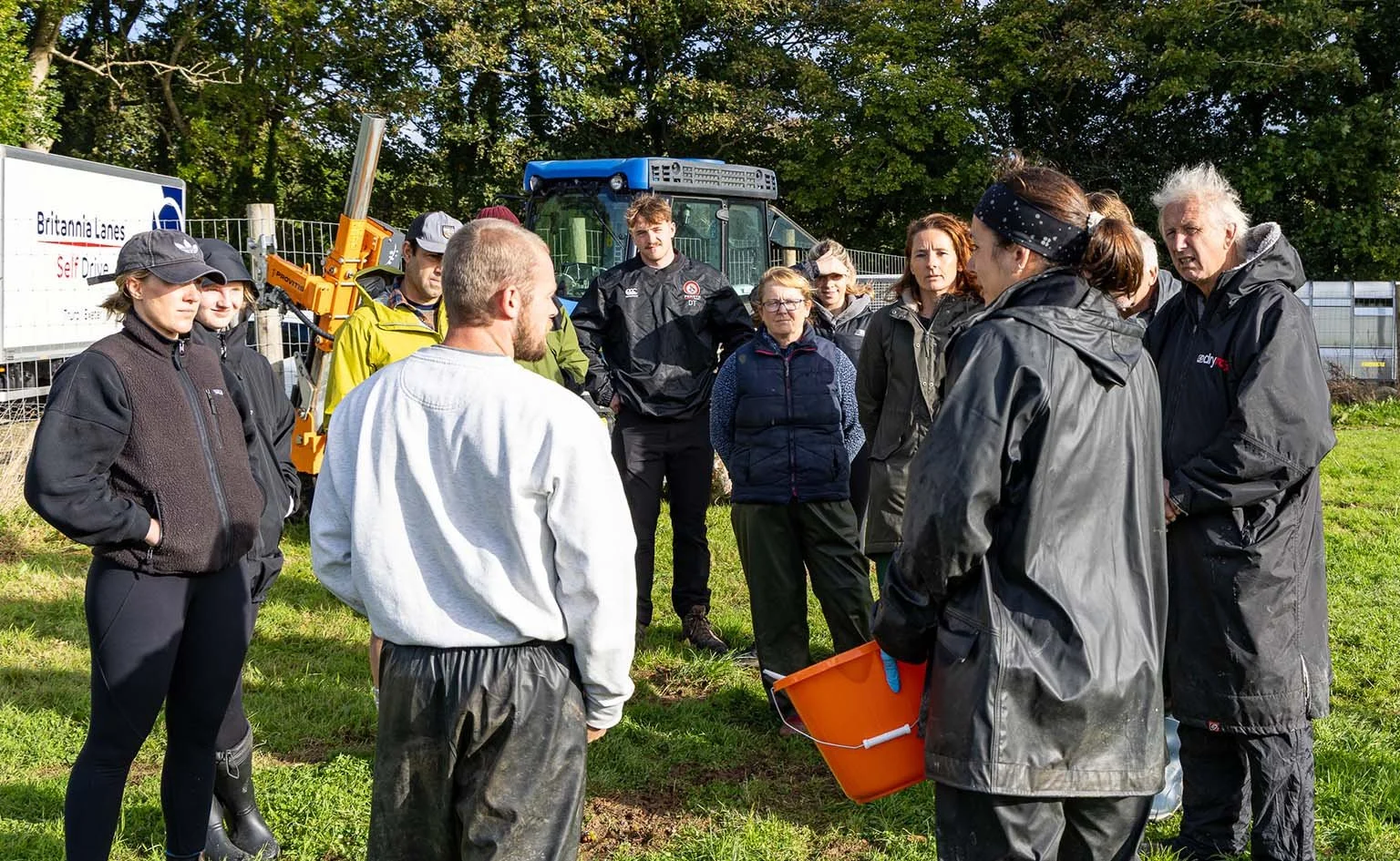 Group of people gathered outdoors in a grassy area, listening to a man speaking. The group is wearing outdoor clothing, and there is a tractor and fencing in the background.