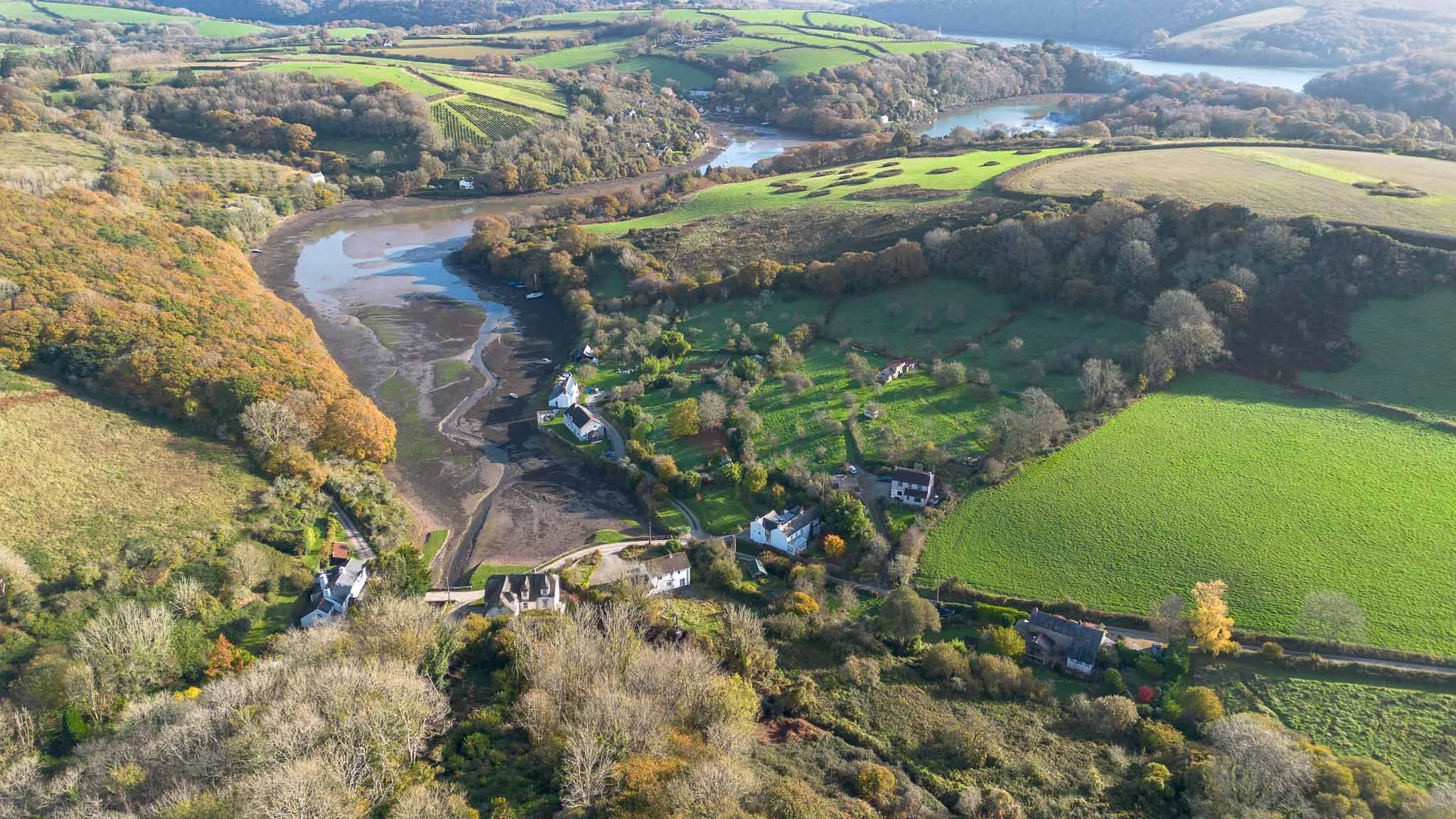 Aerial view of a rural landscape with green fields, trees, a river, and scattered houses.