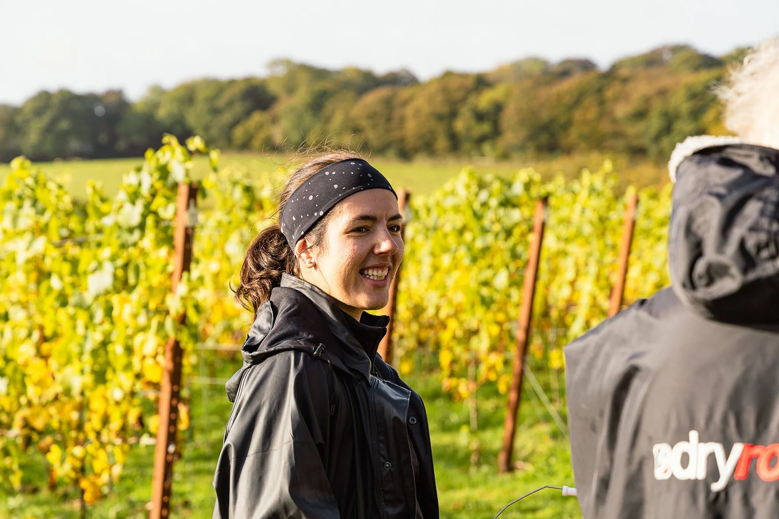 A woman with dark hair tied back, wearing a black headband with white dots and a black jacket, smiling and talking to a person in a black jacket in a vineyard during daytime.