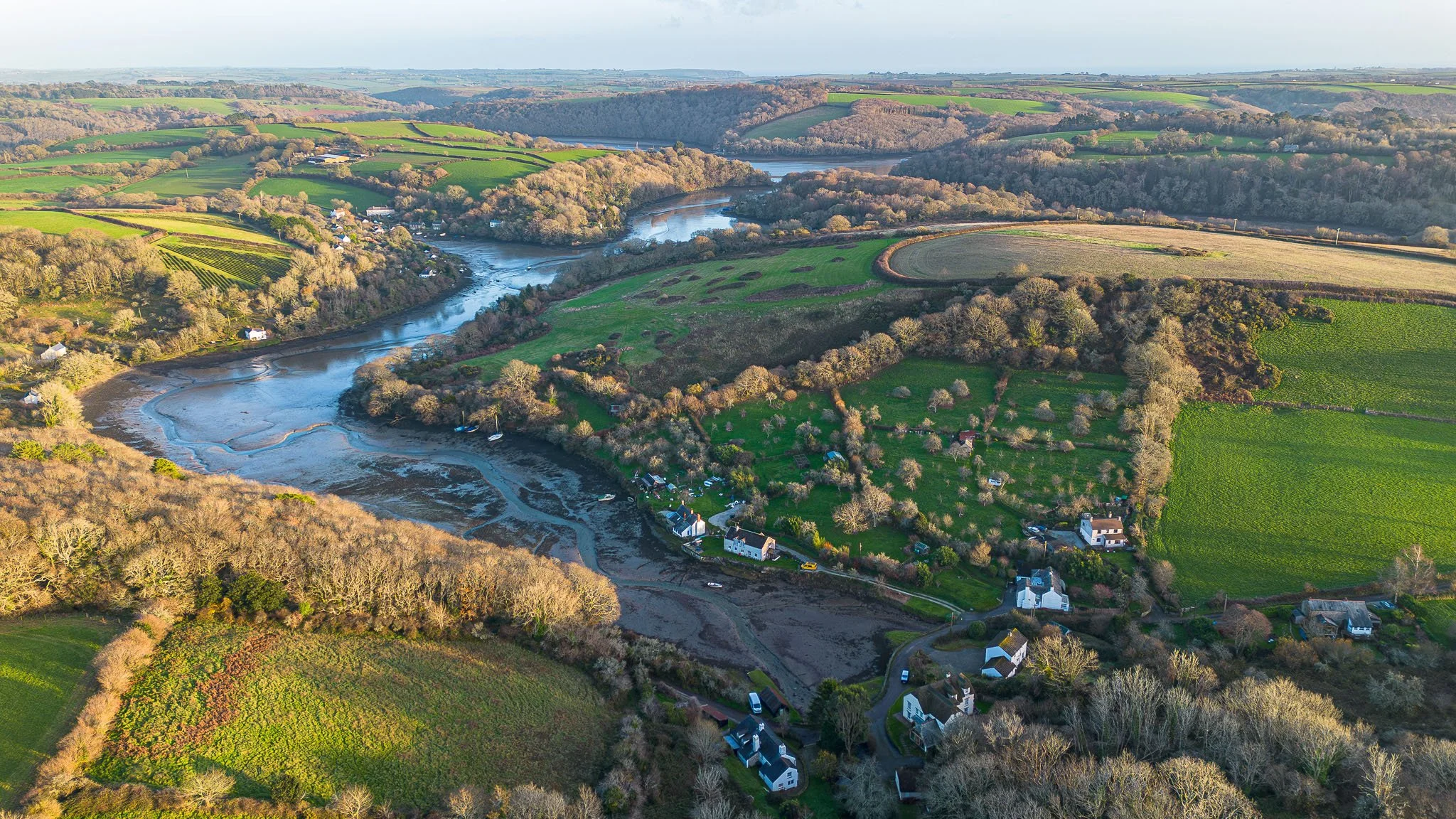 Aerial view of a river winding through a green landscape with houses, trees, and fields in a rural area.