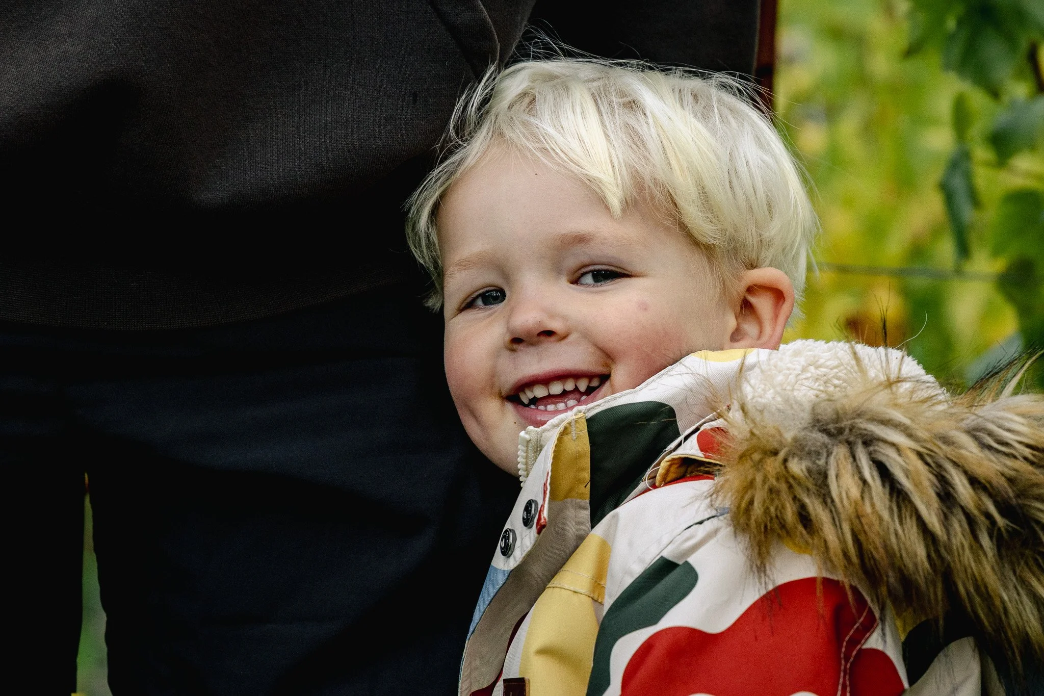 A smiling young boy with blonde hair, wearing a colorful jacket with a faux fur collar, leaning against an adult outside with blurred green foliage in the background.