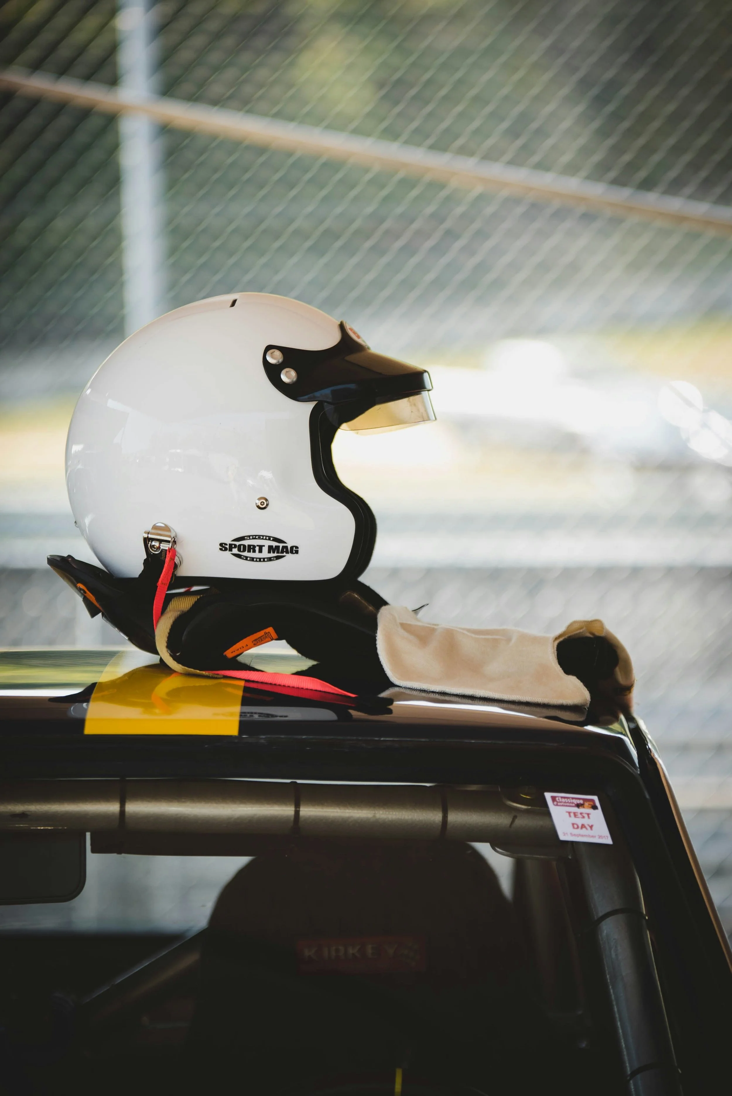 A white racing helmet with black visor and straps resting on a black car's roof, with a gray cloth and sticker reading 'TEST DAY' nearby.