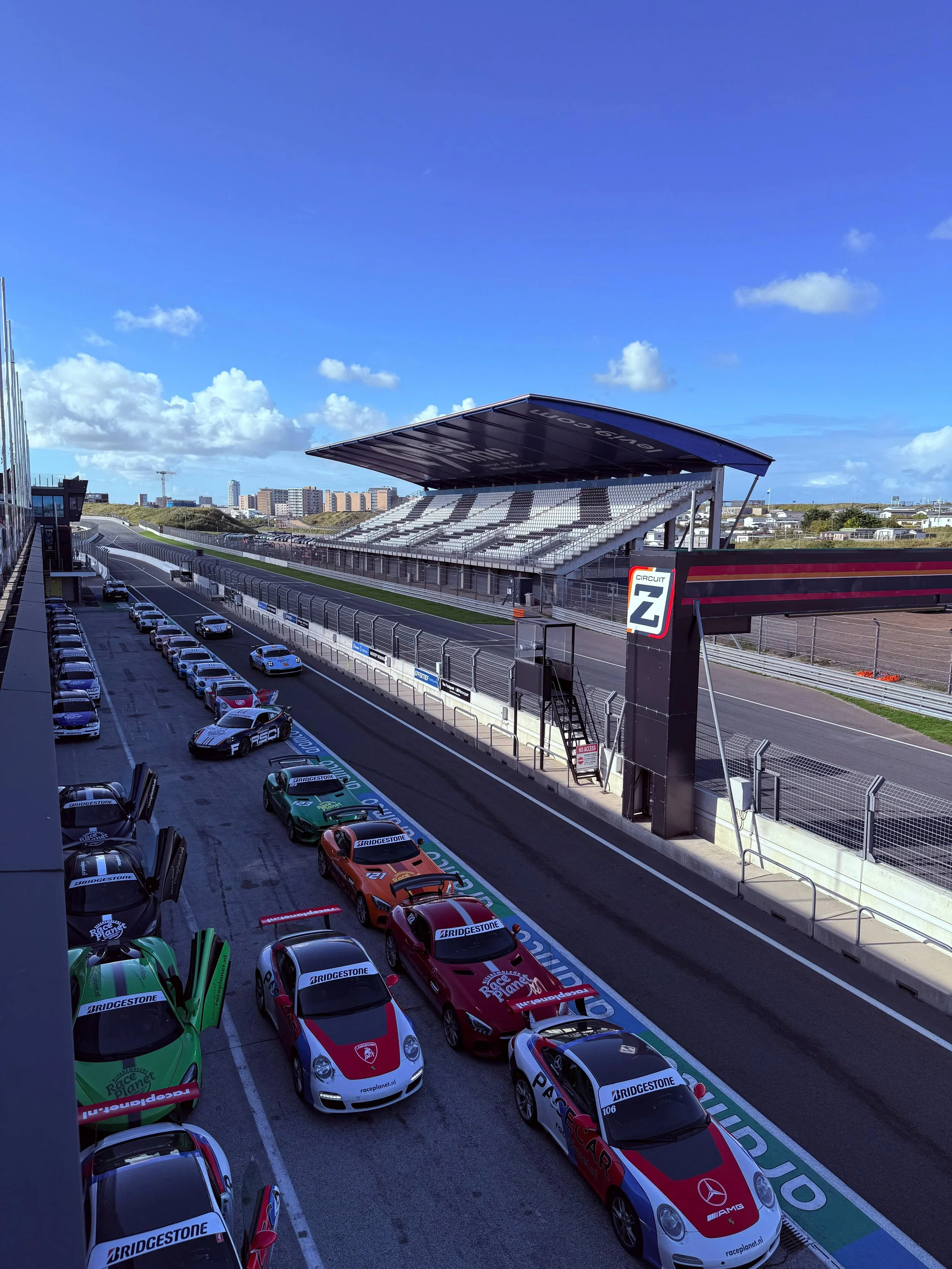 Race cars parked in the pit lane at a race track, with grandstands and a blue sky in the background.