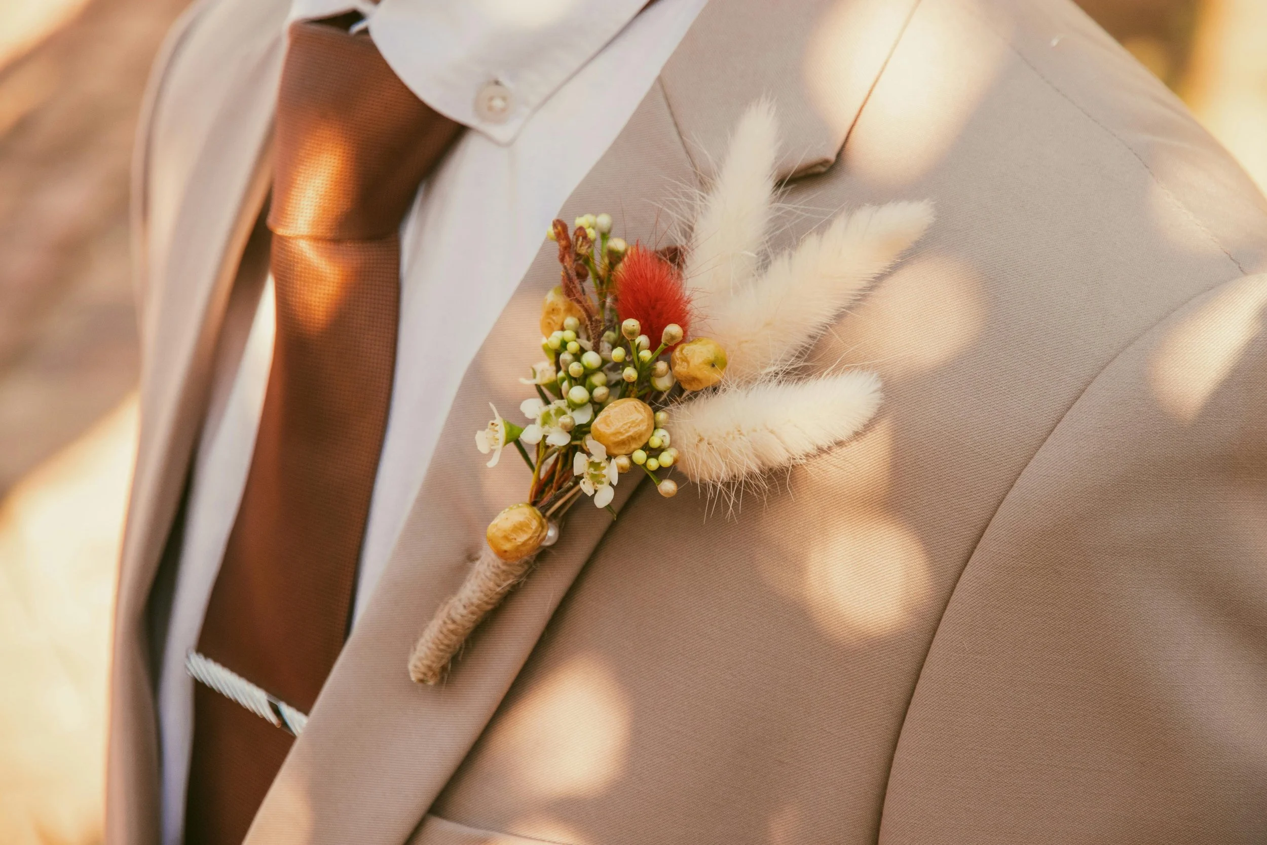 A man wearing a beige suit with a boutonniere made of white, yellow, and red feathers, small white flowers, and yellow berries pinned to his lapel.