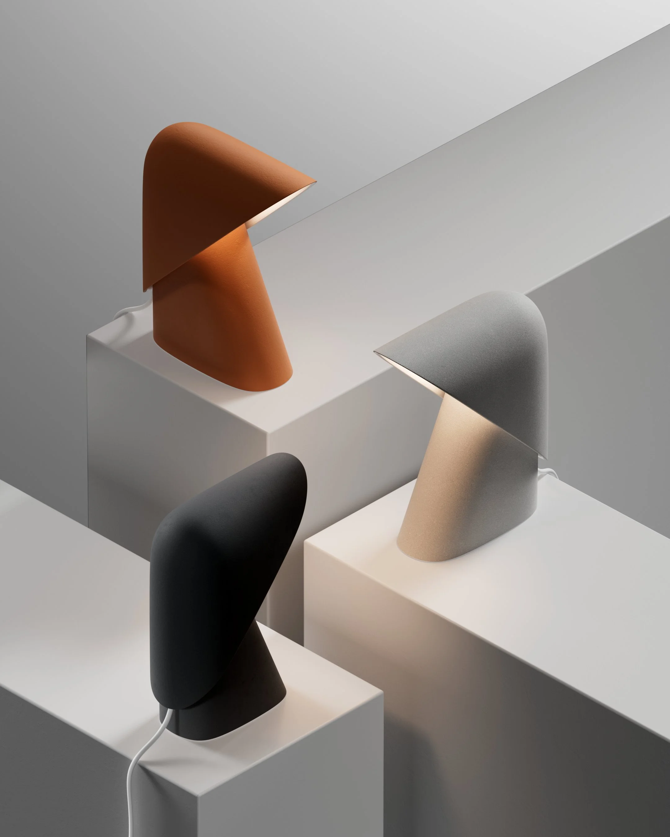 Tre lampade da tavolo moderne di diversi colori, posizionate su superfici bianche, con illuminazione soffusa in uno sfondo grigio chiaro.