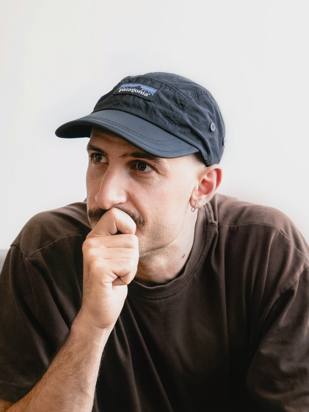 Uomo che indossa un cappello Patagonia e una maglietta marrone, pensieroso, con la mano sulla bocca.