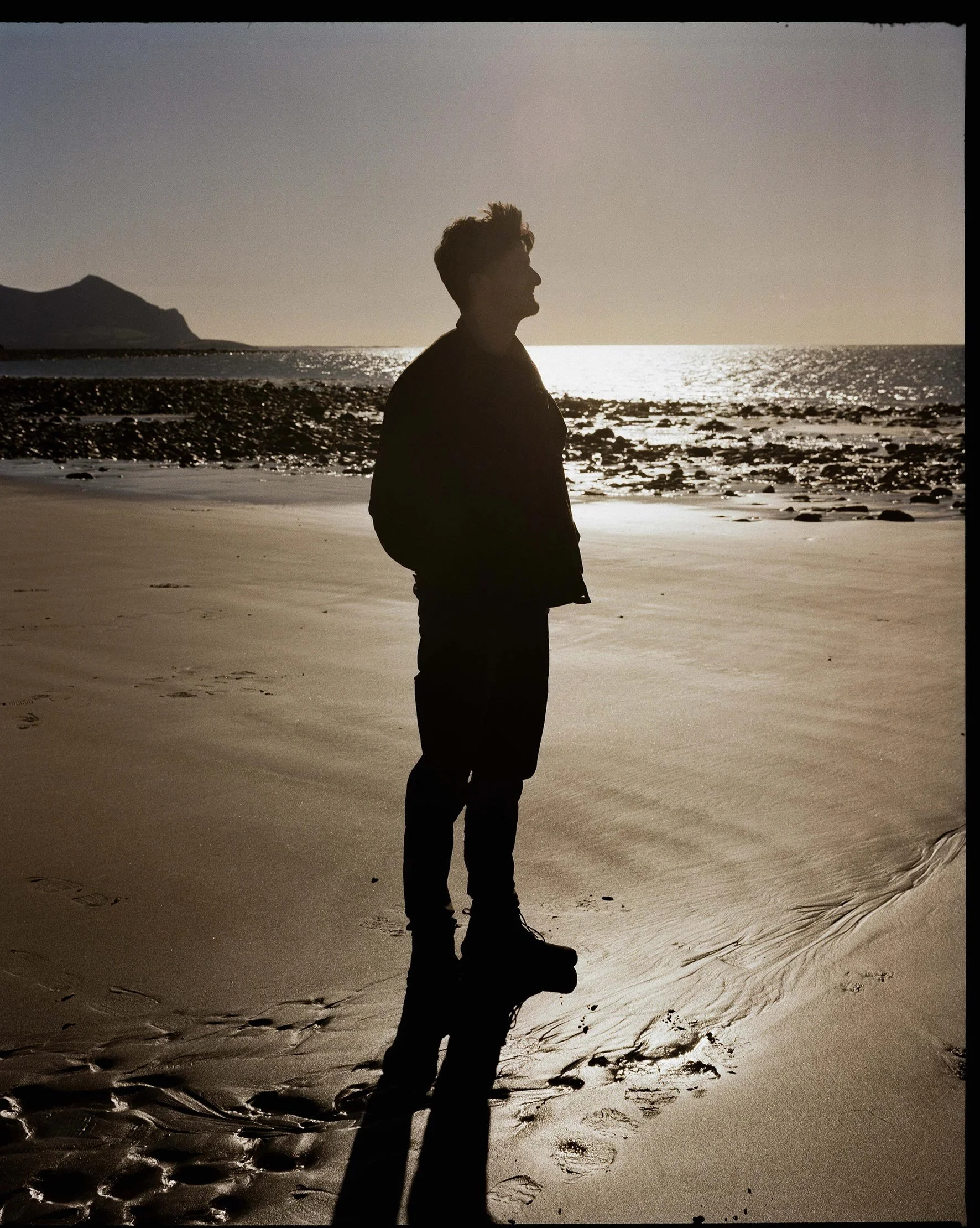 A man stands on a sandy beach at sunset, silouetted by the sun