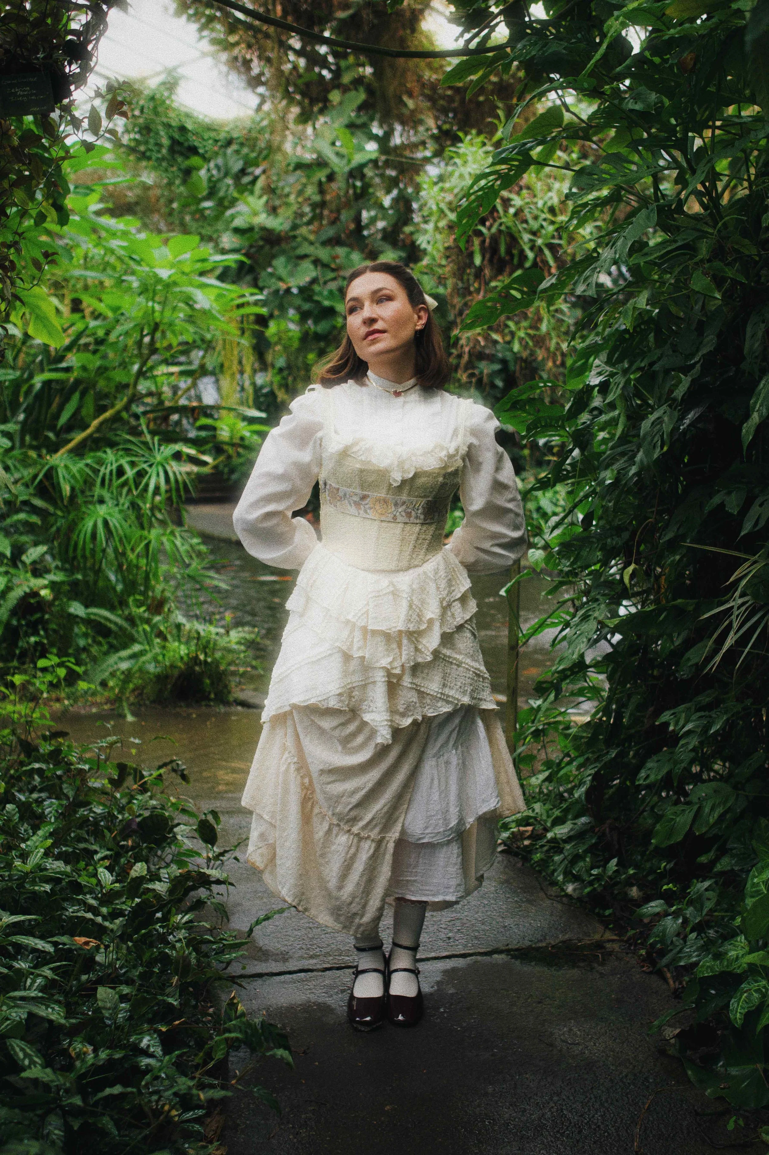 Mared wearing a long white ruffled dress looks up to the sky. She is surrounded by lush green foliage