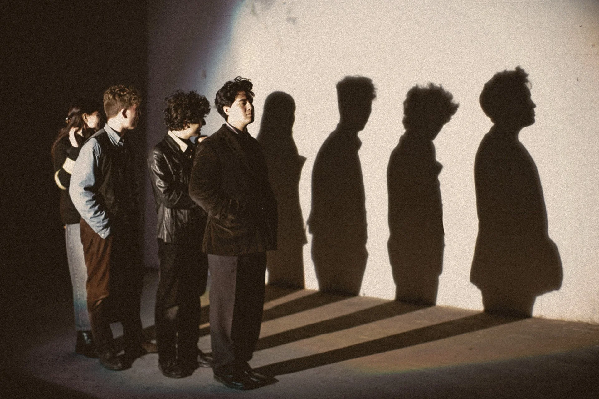 Four band members of SLATE stand in a row with their shadows reflected against a pale wall behind them