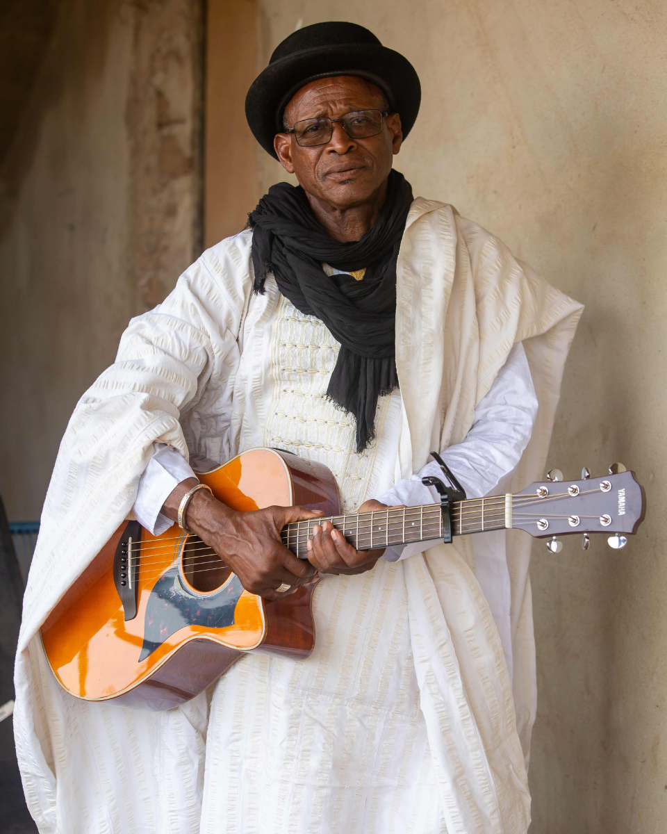 Afel Bocoum dressed in traditional white attire, wearing a black hat, glasses, and a black scarf, playing an acoustic guitar.