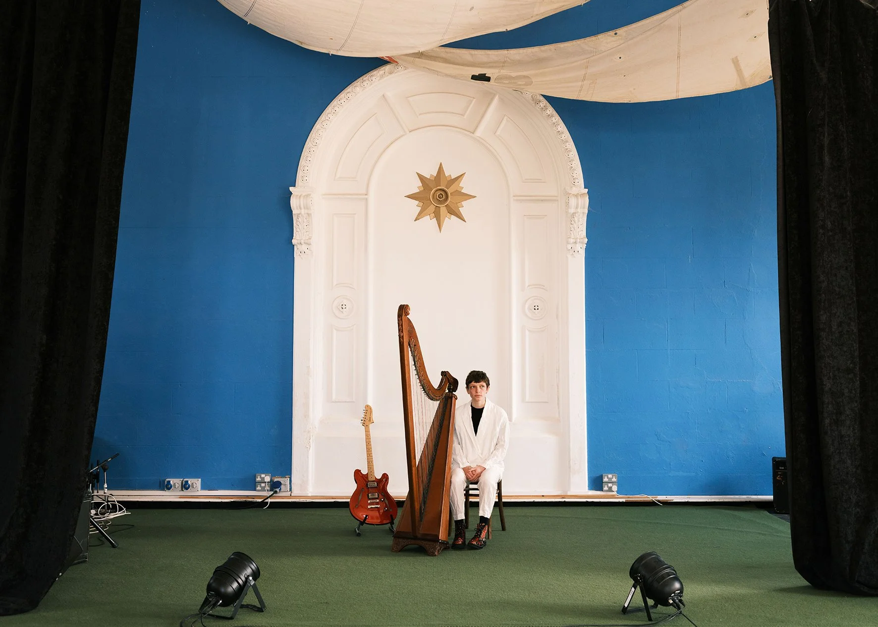 Cerys Hafana in a white suit sits with her harp and guitar in front of a large white door with a gold star motif