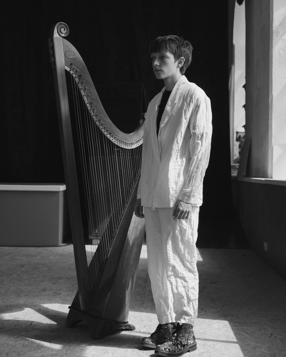 Cerys Hafana standing next to a large harp in a room with natural light, wearing a light-colored jacket and pants, with dark hair.