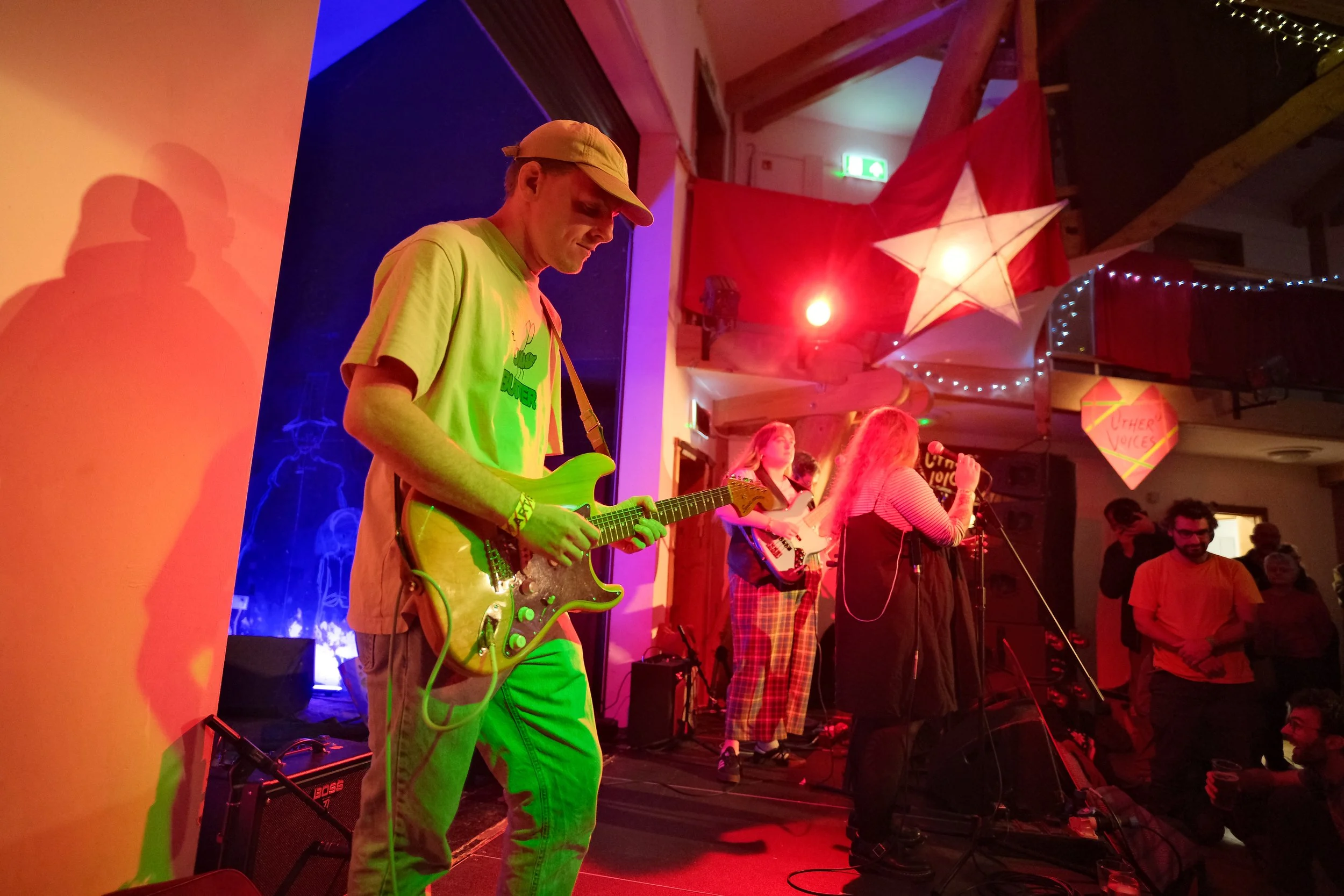 A band performing on stage with a guitarist, a bassist, and a vocalist in a cozy, decorated indoor venue with colorful lighting, star and heart decorations, and string lights in the background.